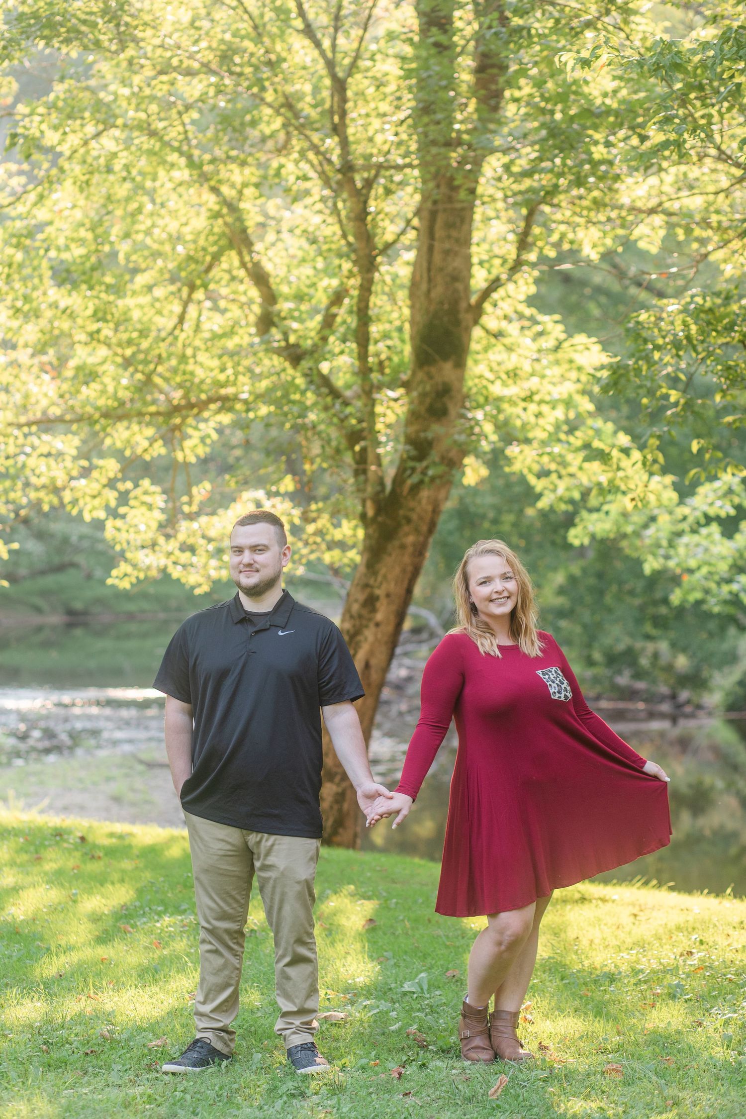 A couple poses playfully in a sun-dappled park setting with golden autumn trees.