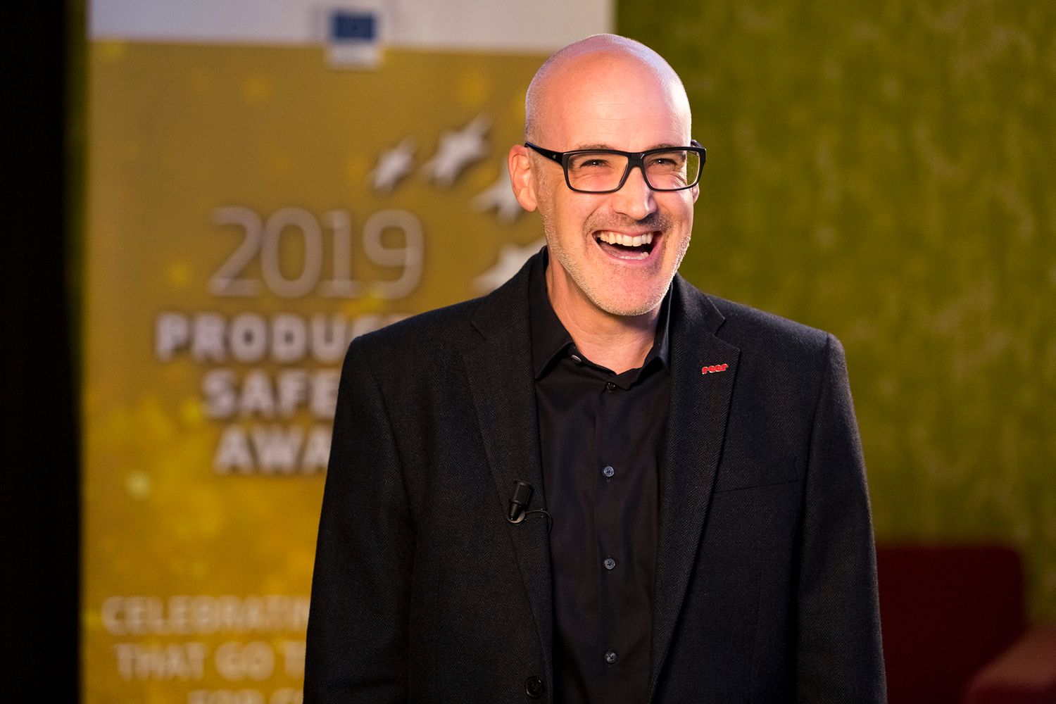 Speaker presenting at 2019 Product Safety Awards against yellow backdrop.