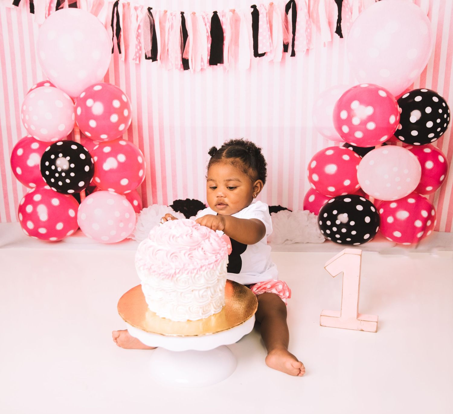 Baby in white outfit enjoying their first birthday cake surrounded by pink and black polka dot balloons.