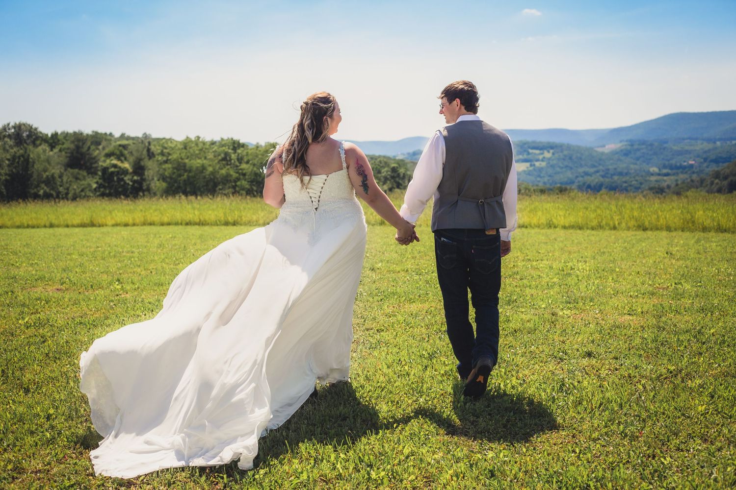 Couple walking hand in hand across sunny meadow with flowing white wedding dress.