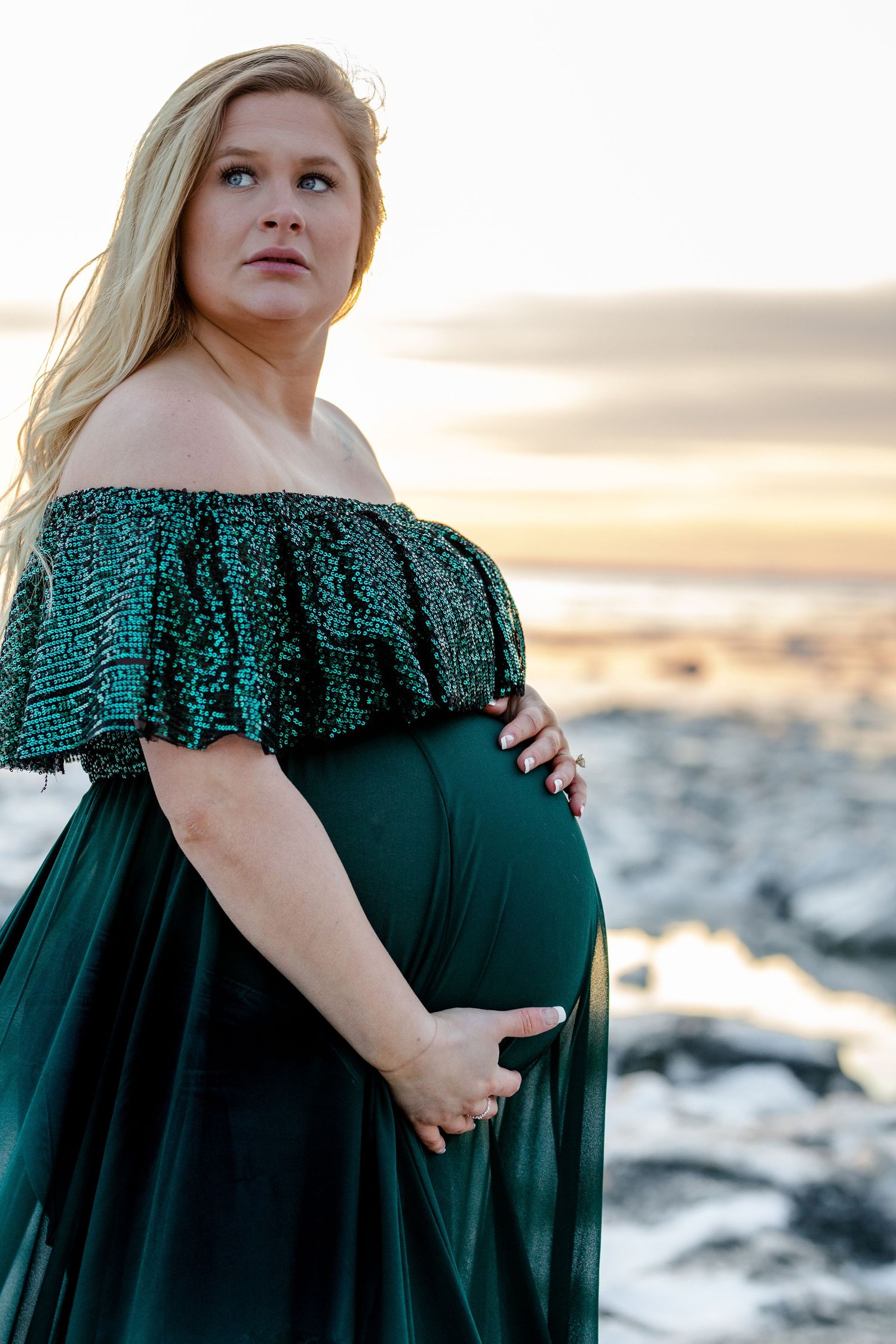 Maternity photo at sunset featuring emerald green off-shoulder dress on rocky beach.