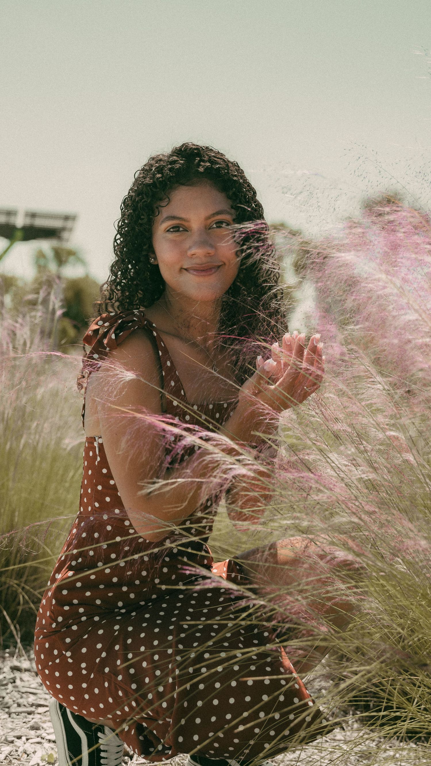 Woman in polka dot dress poses joyfully among flowing ornamental grasses.