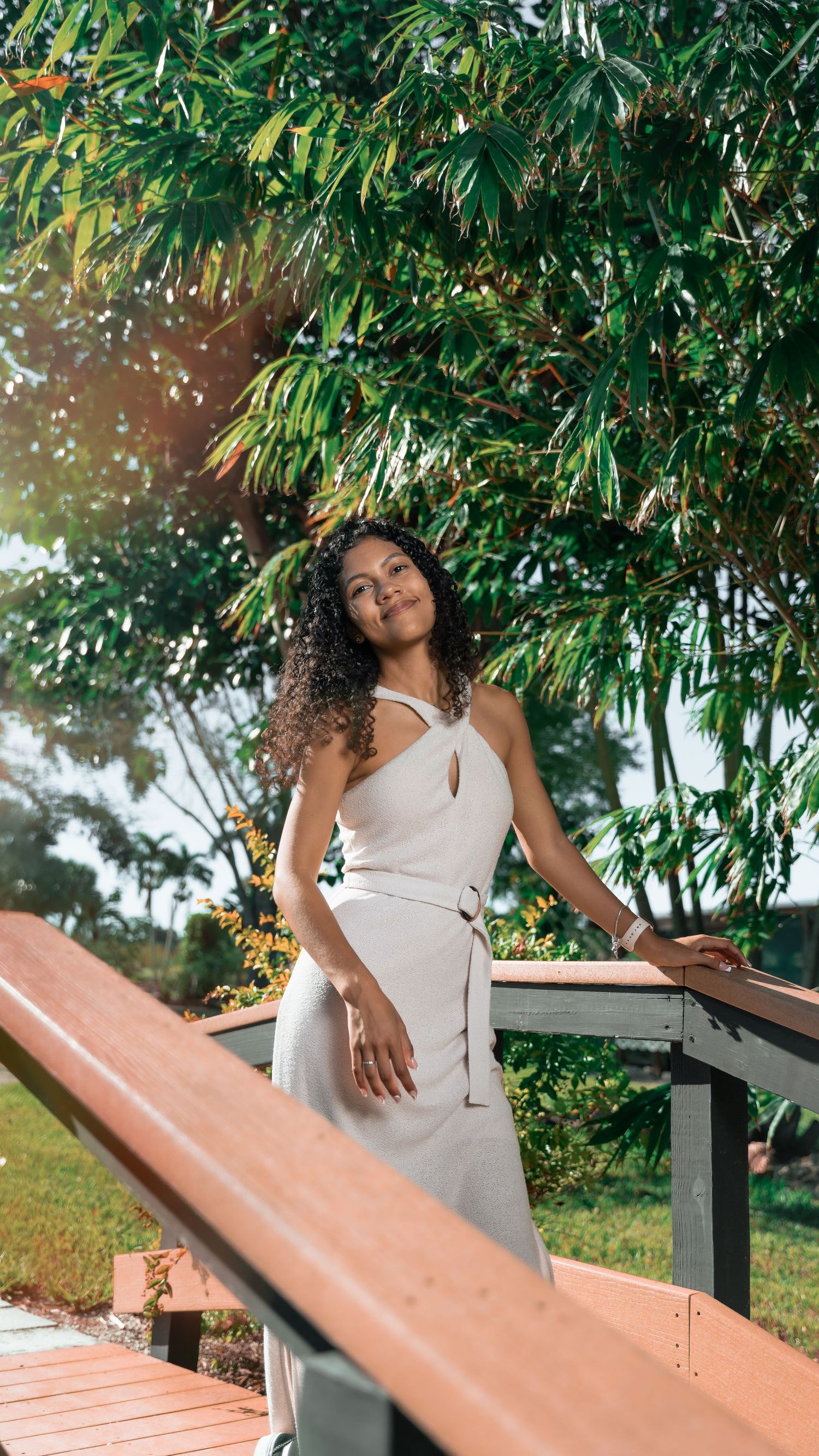 Person in an elegant white dress poses on a wooden deck beneath lush green tropical foliage on a sunny day.
