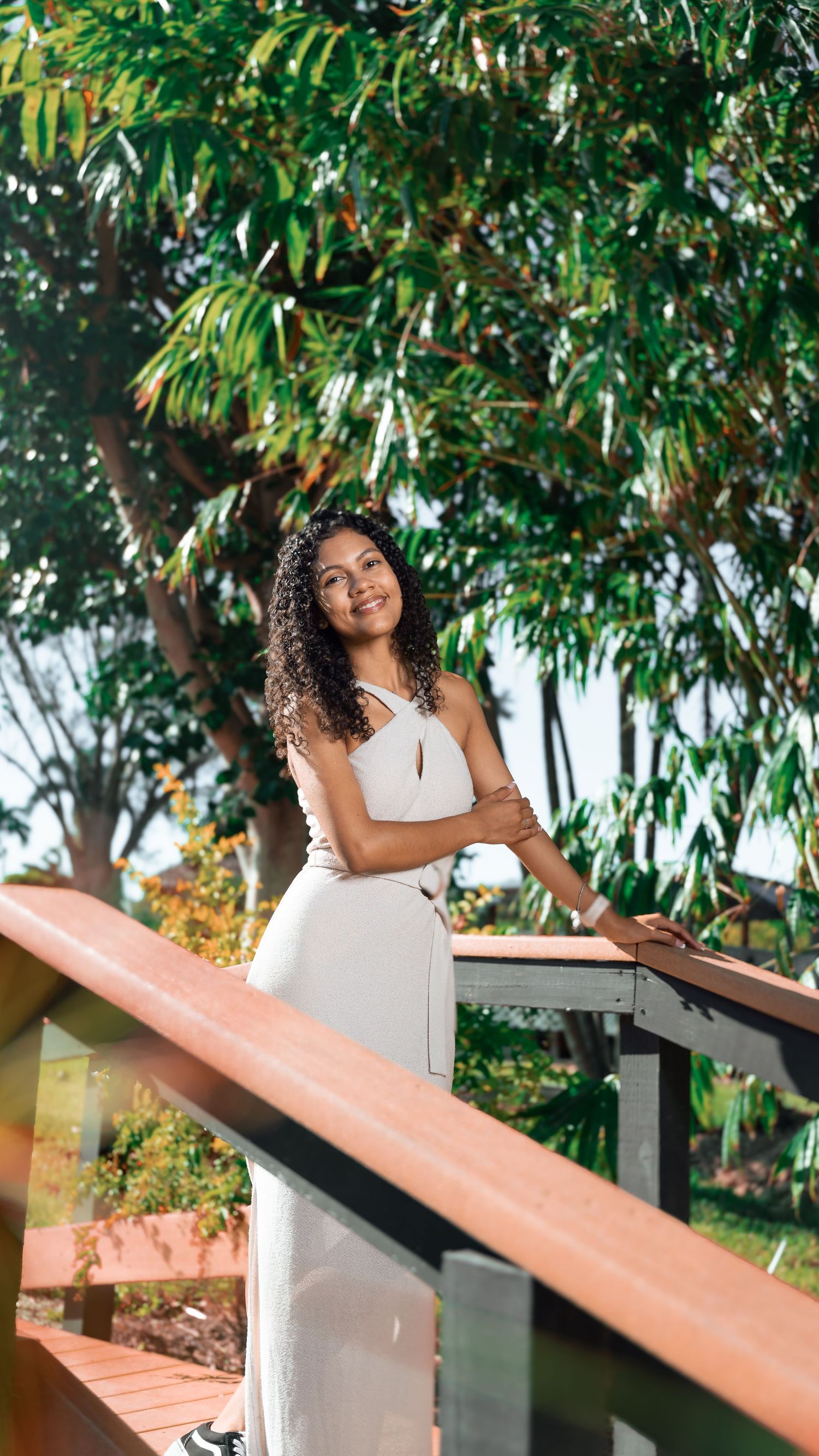 Woman in white dress poses on wooden deck with tropical foliage backdrop.