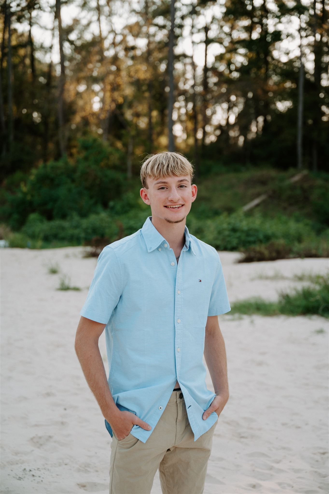 Person in light blue button-down shirt and khaki pants poses casually on sandy beach.