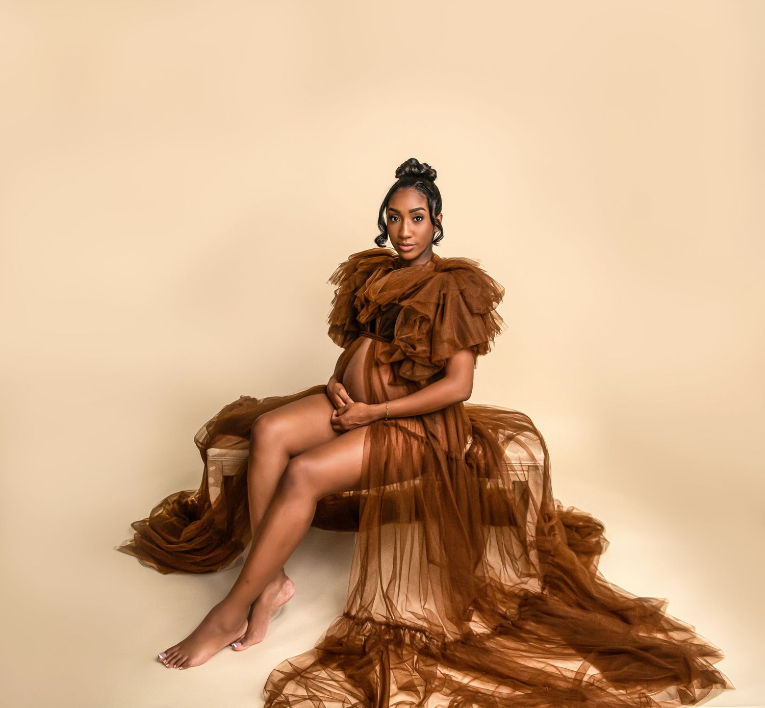 A glamorous maternity portrait featuring a brown ruffled gown against a beige backdrop.