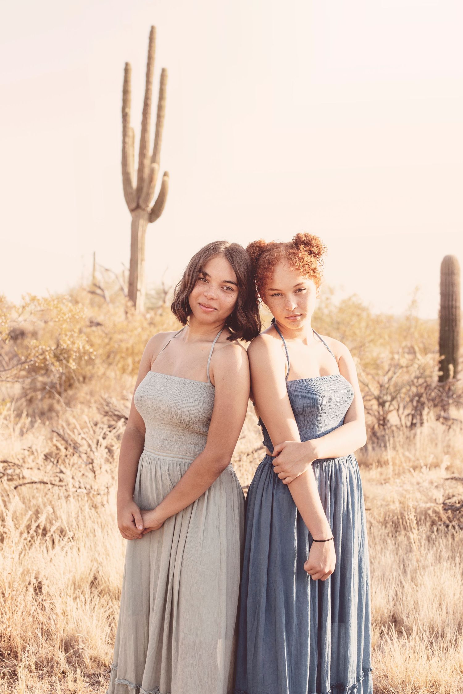 Two women in flowing dresses pose together in desert landscape with saguaro cactus at sunset.