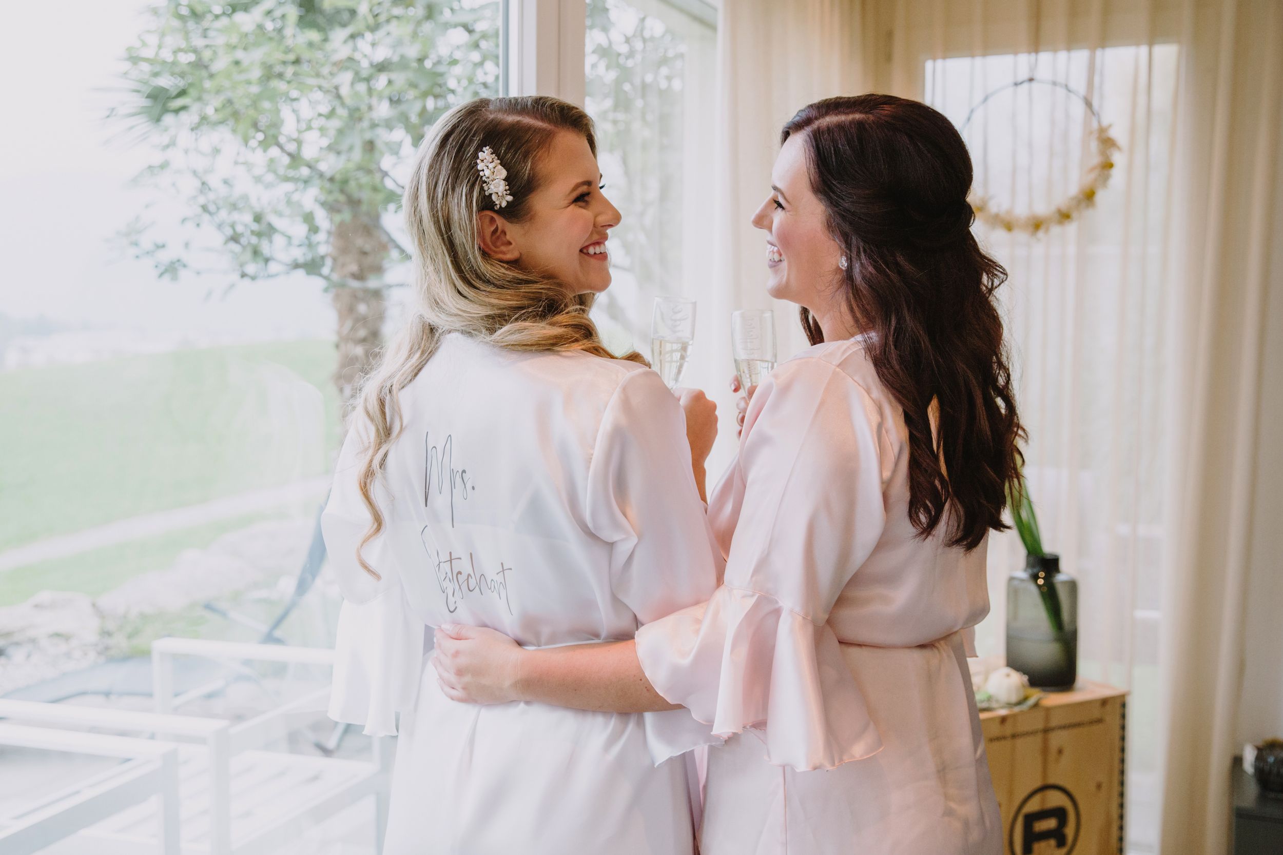 Bride and bridesmaid in matching white silk robes smiling together during wedding morning prep.