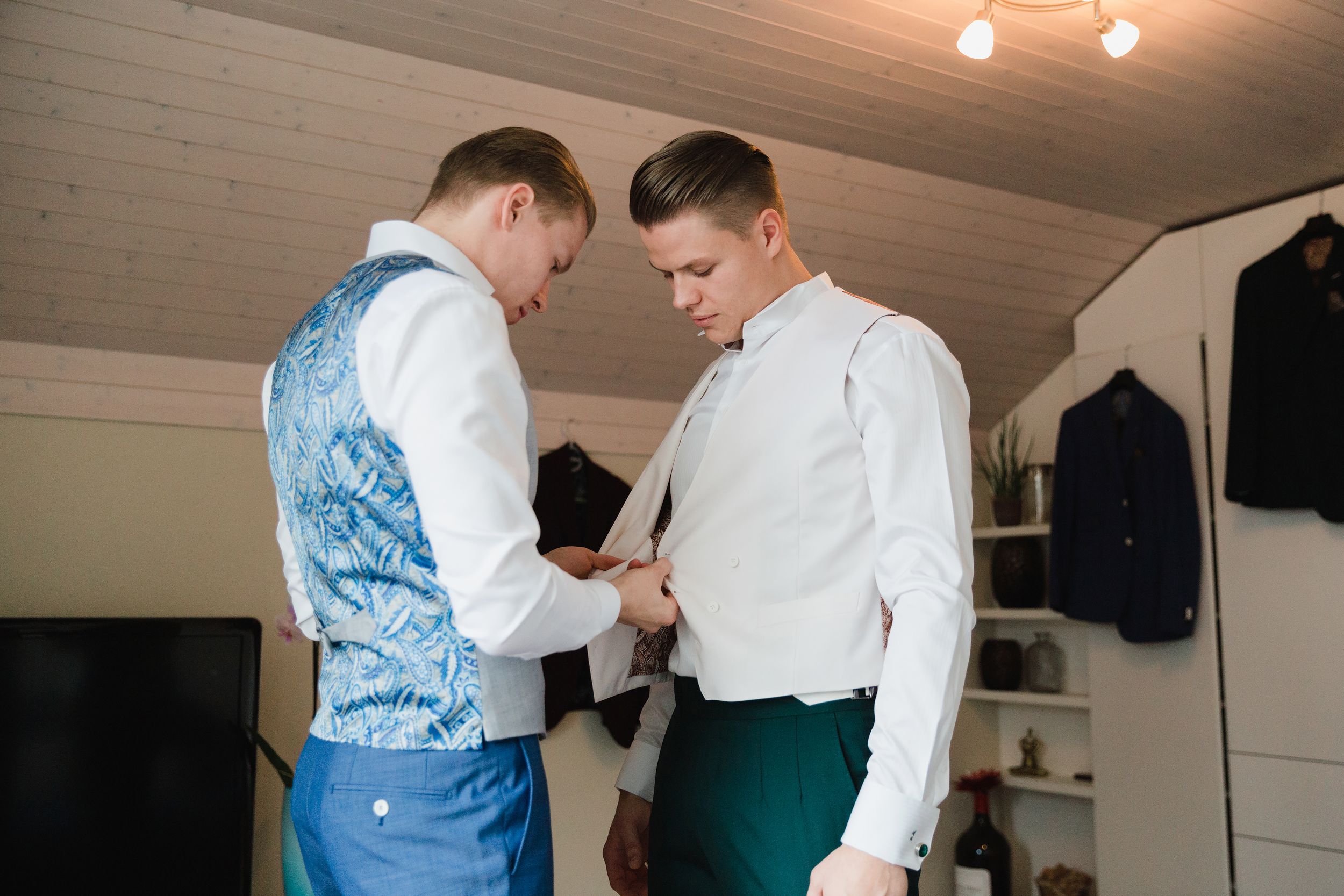 Close-up of a groomsman in a floral vest helping the groom fasten his white dress shirt.