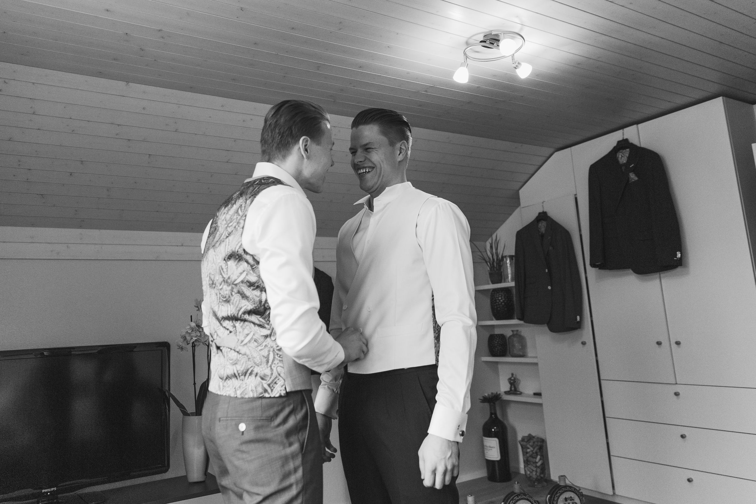 Black and white candid of a groom and groomsman talking while getting ready in a bright dressing room.