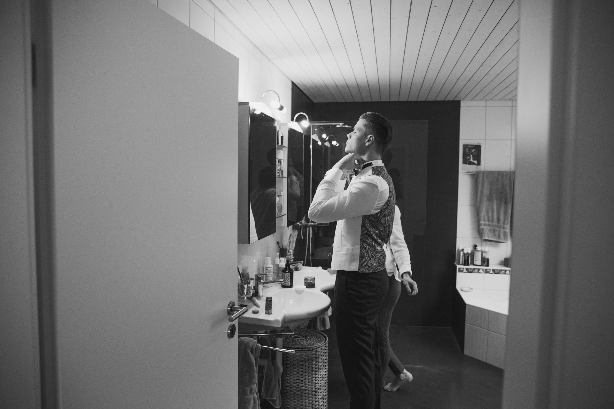 Black and white candid of a groom adjusting his collar in a bathroom mirror during wedding morning prep.
