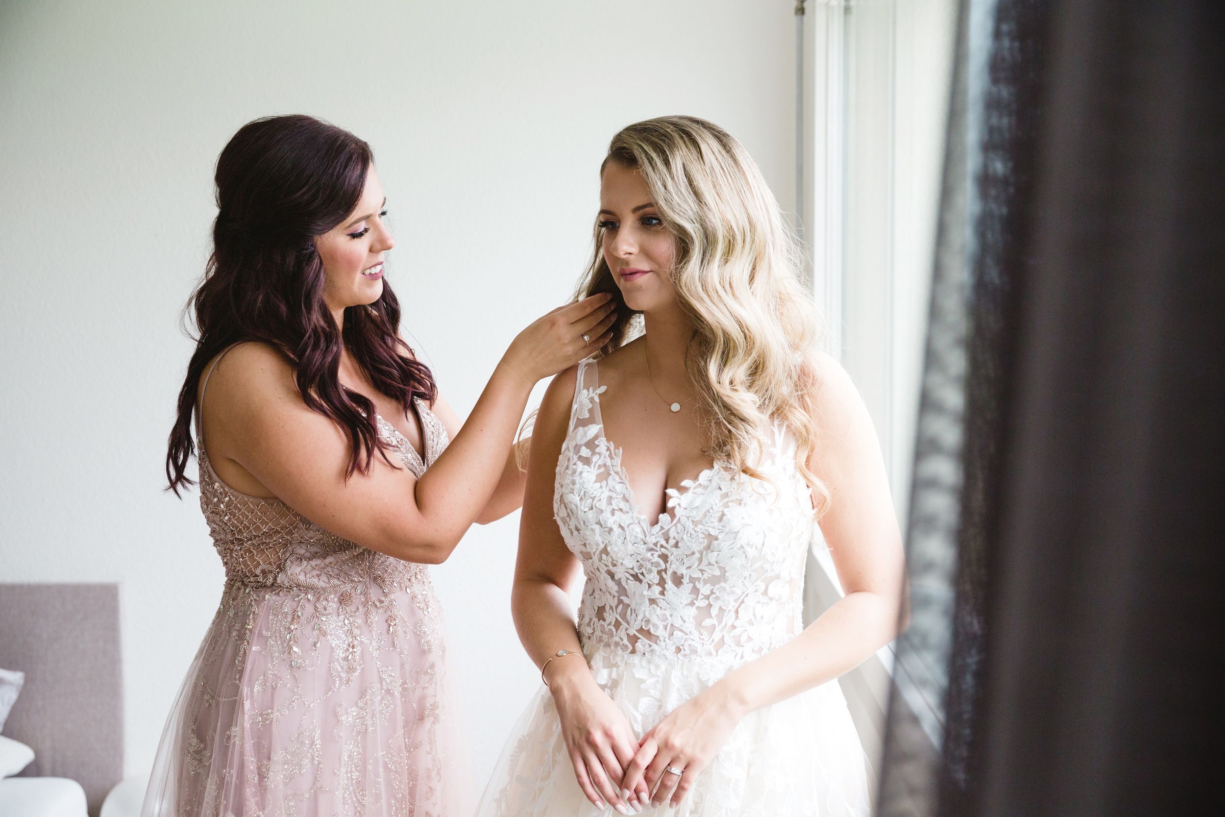 Bridesmaid in a pink dress helping the bride fasten her white lace wedding gown.