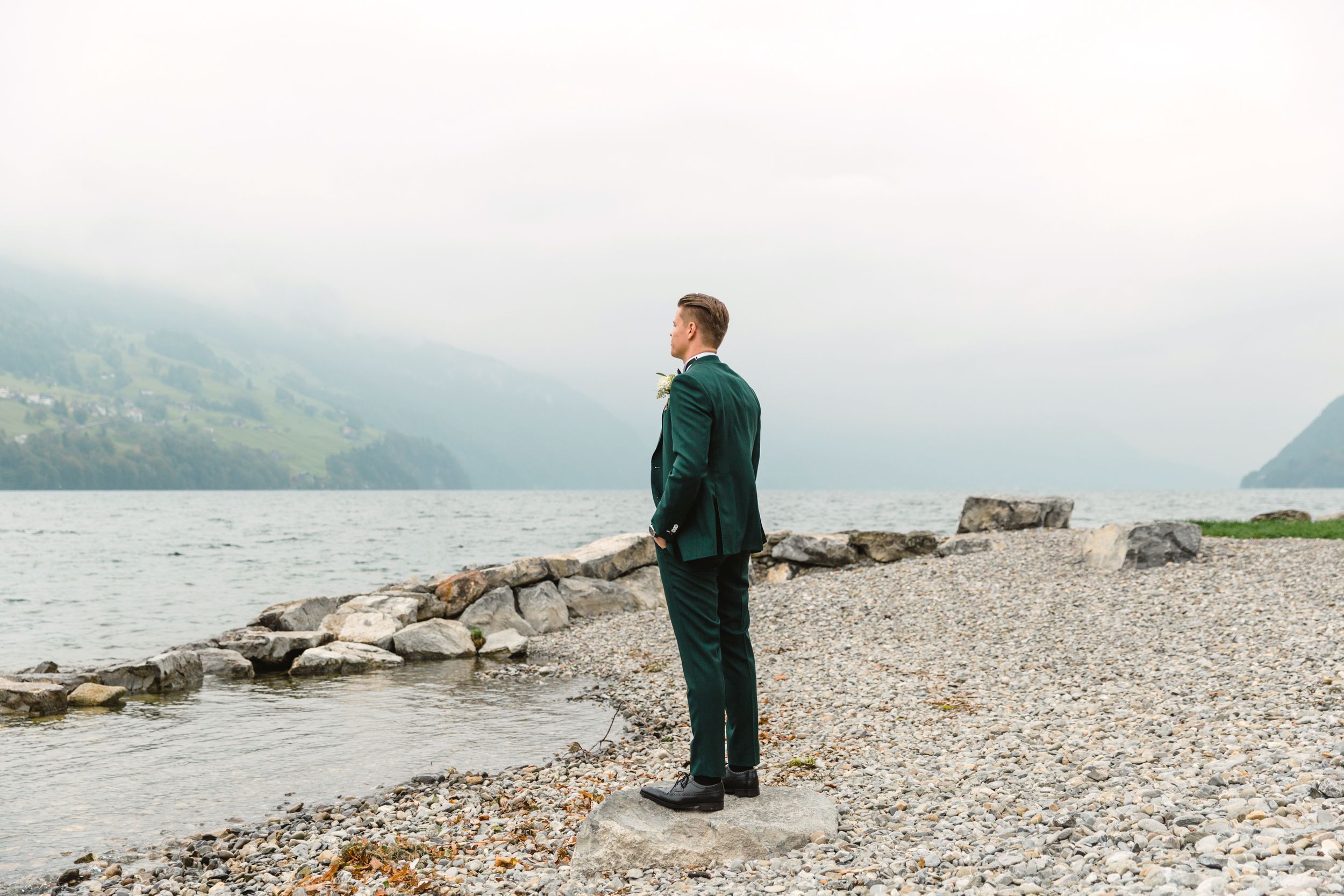 Groom in a dark green suit standing on a rocky lakeshore, looking out at the misty Swiss mountains.