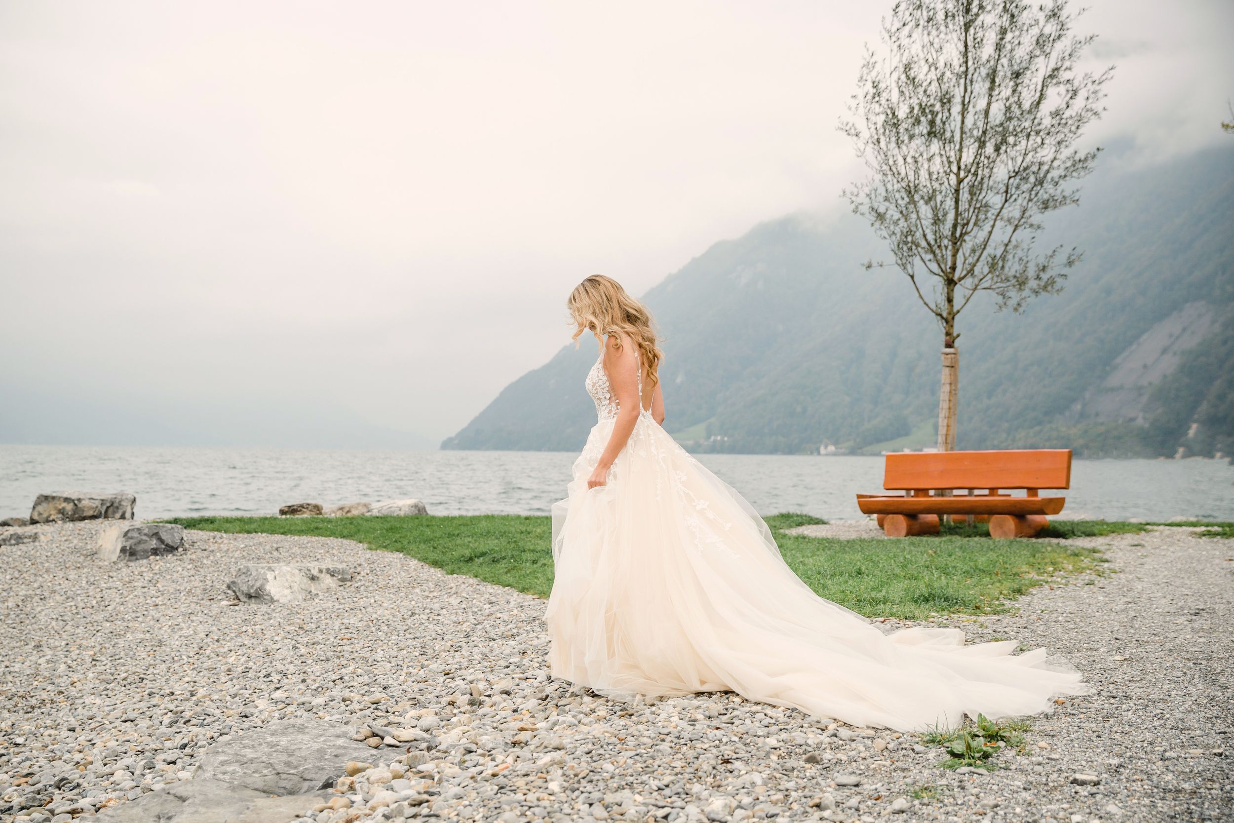 A bride in a white lace gown standing on a stone ledge by a lake with a solitary tree and mountains.