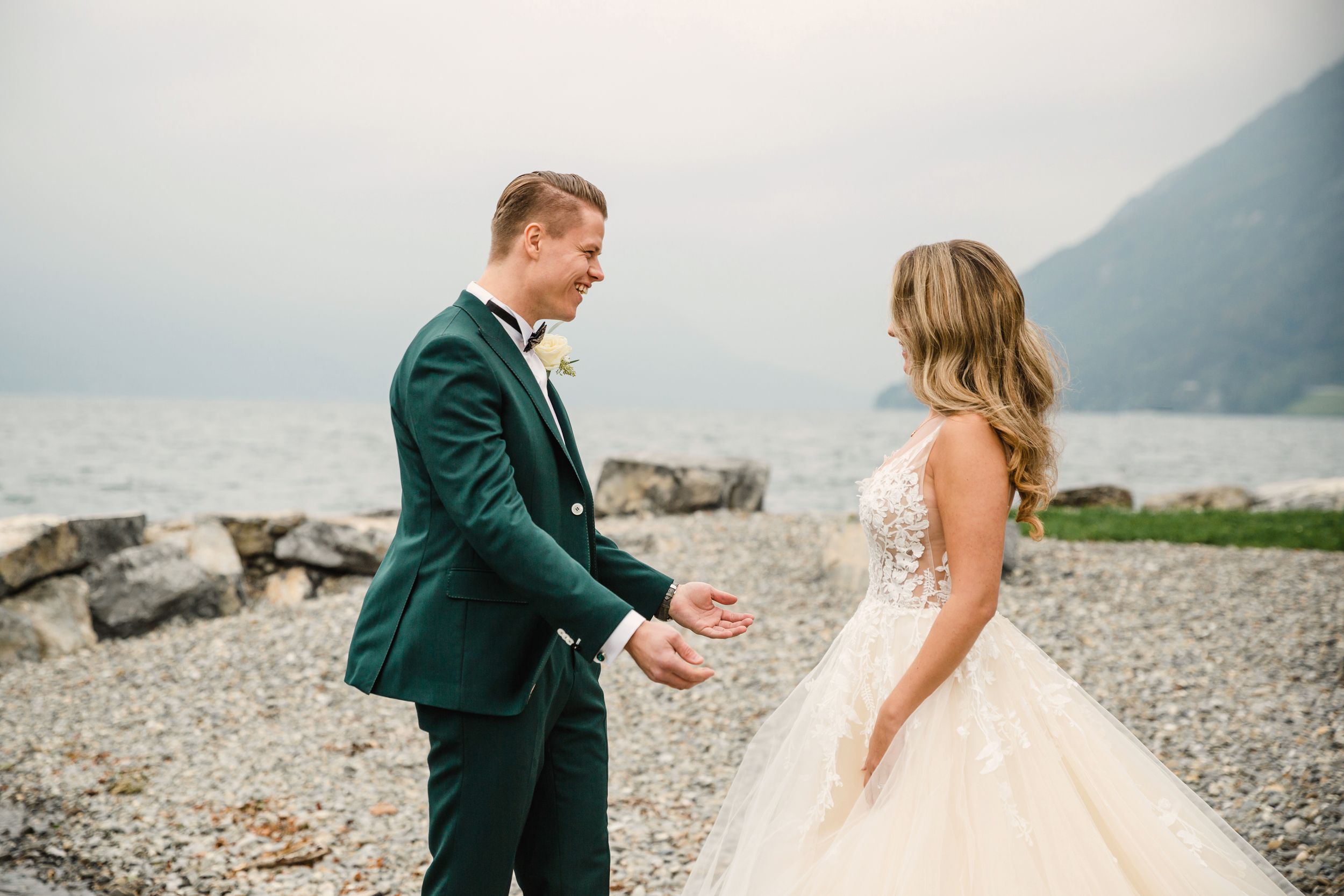Groom in a dark green suit smiling at his bride on a rocky lakeside path in Brunnen.