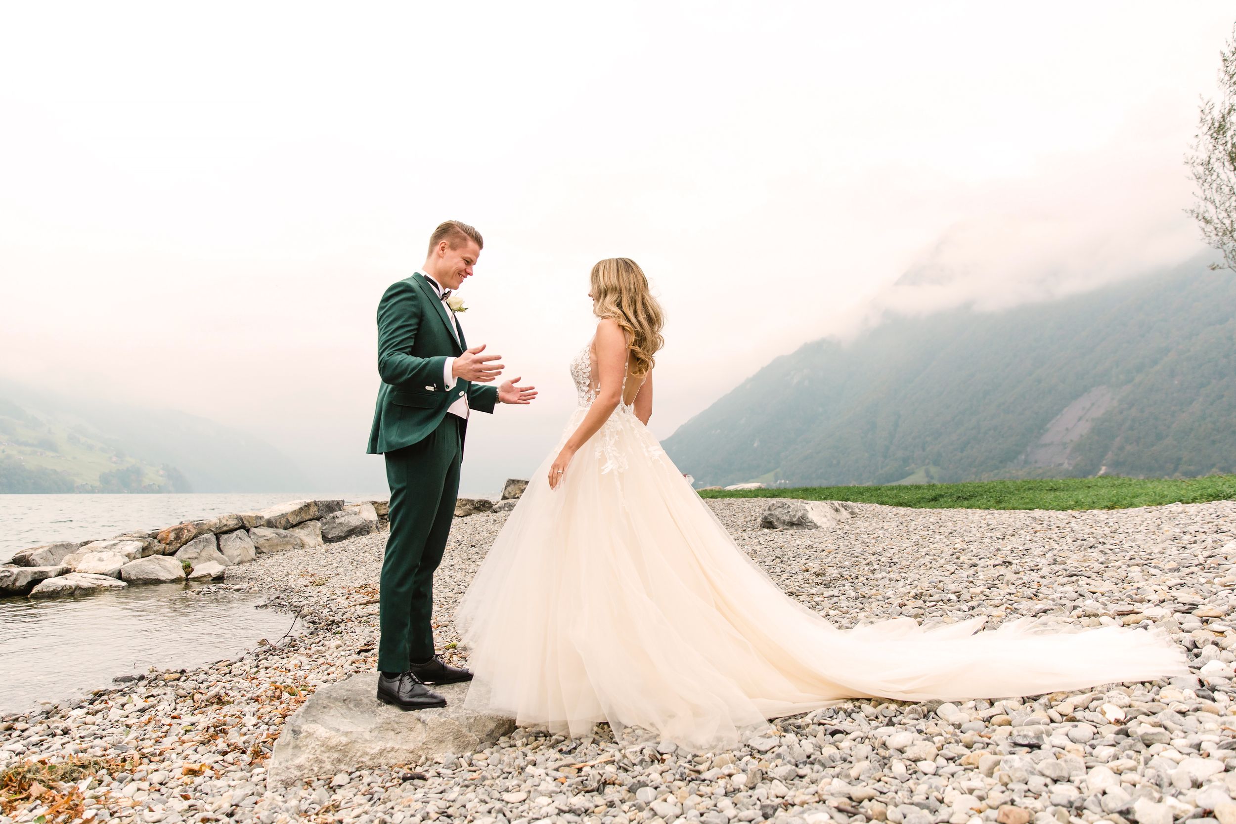 Bride and groom standing together on a pebble beach with a vast mountain lake in the background.