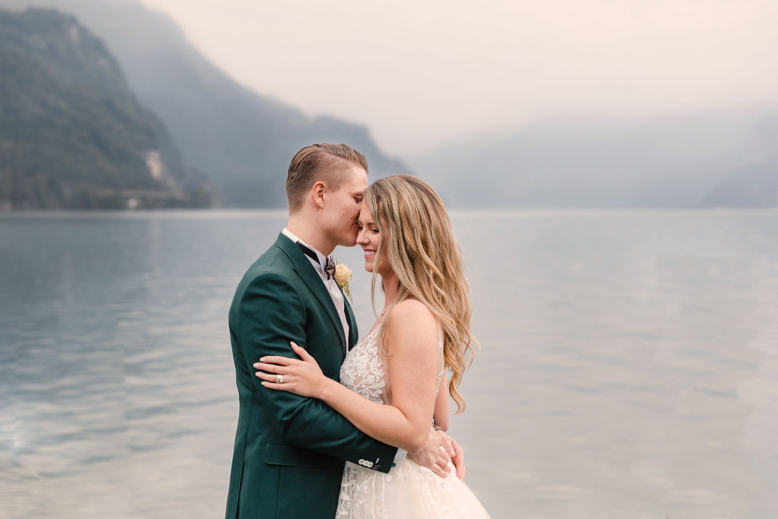 Romantic wedding portrait of a bride and groom embracing by a scenic lake with misty mountains in the background.