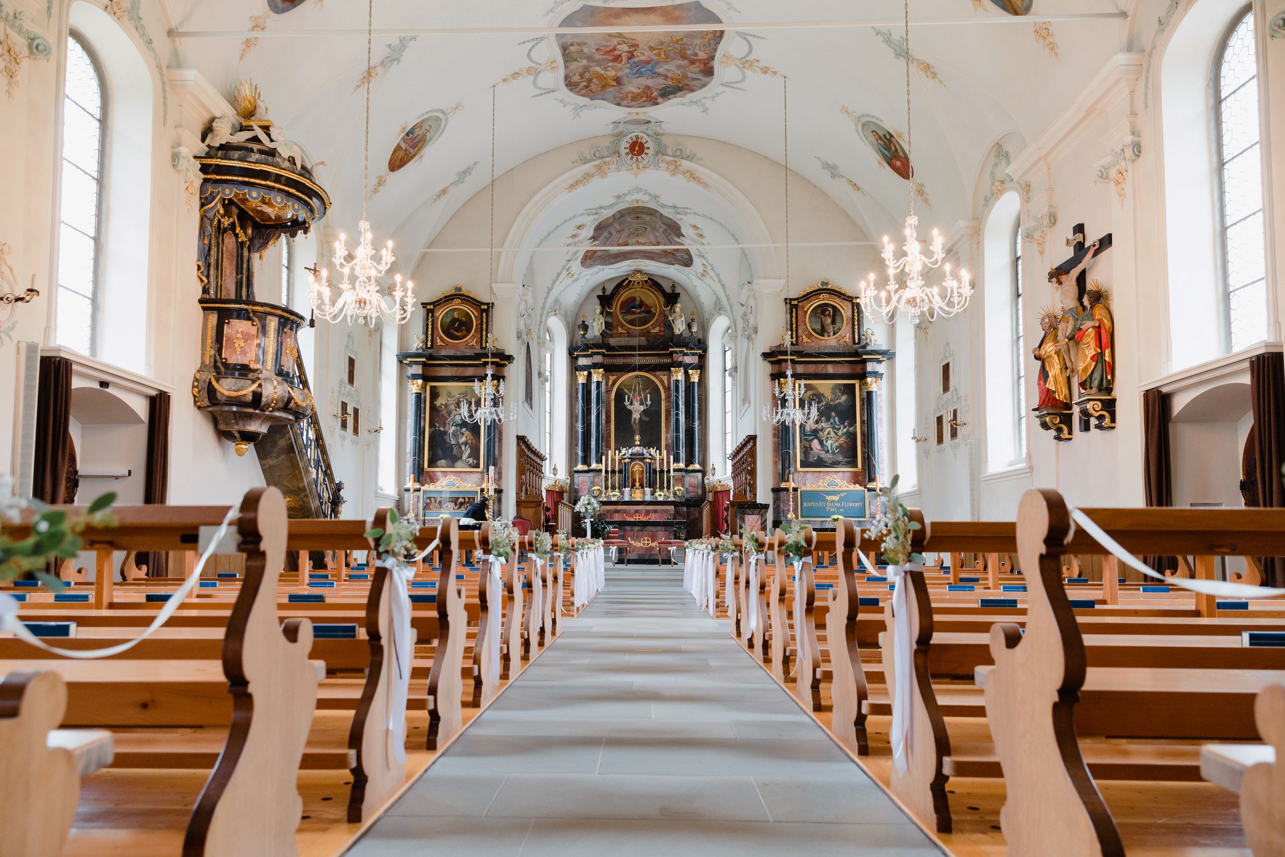 Wide interior view of an ornate, bright church nave with wooden pews and a golden Baroque altar.