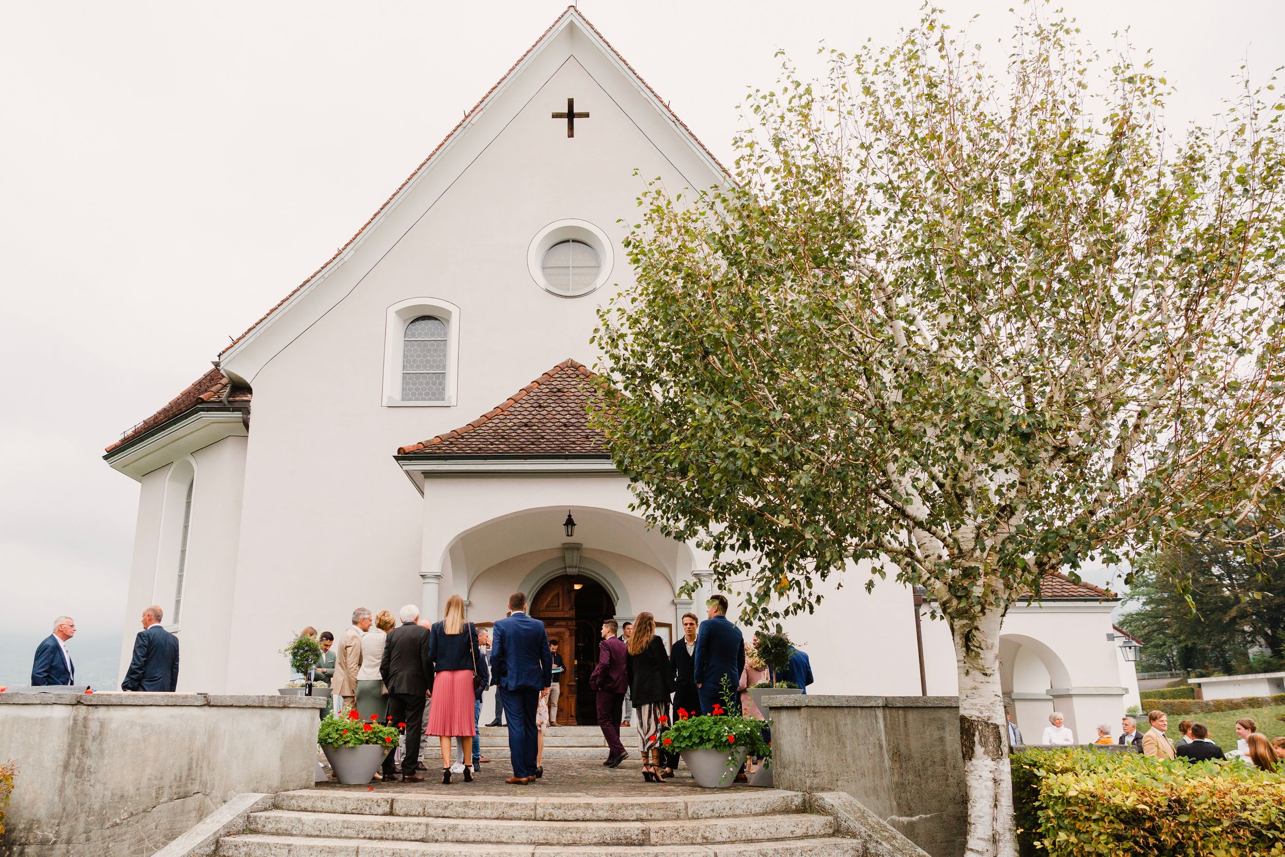 Exterior shot of guests gathering in front of a white historic church with a red-tiled roof for a wedding.