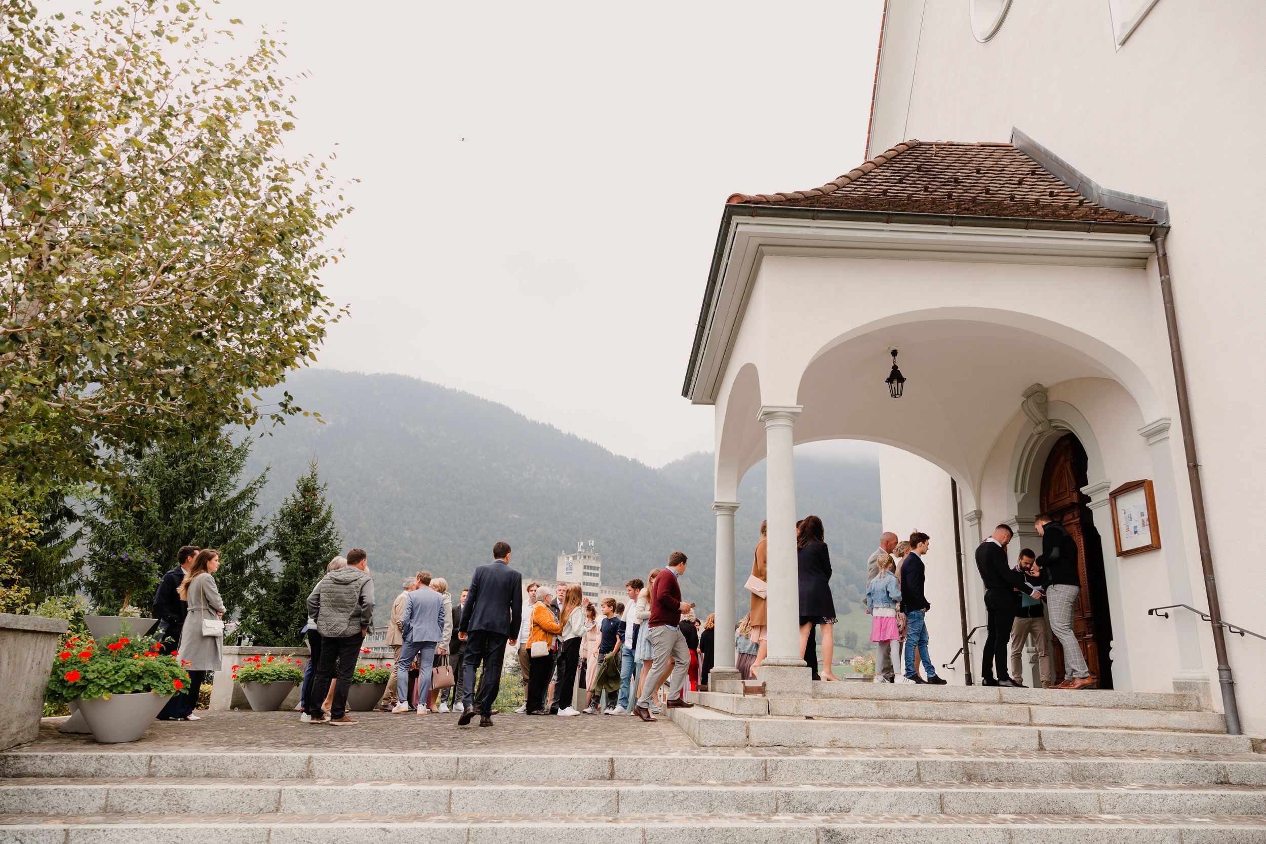 Wedding guests gathered outside a white chapel or church entrance in a scenic mountain setting.