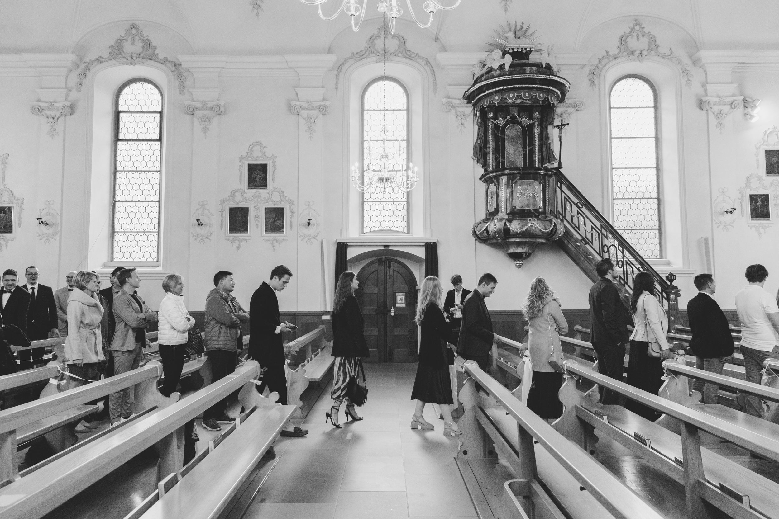 Black and white photo of wedding guests seated in the wooden pews of a historic, sunlit church.