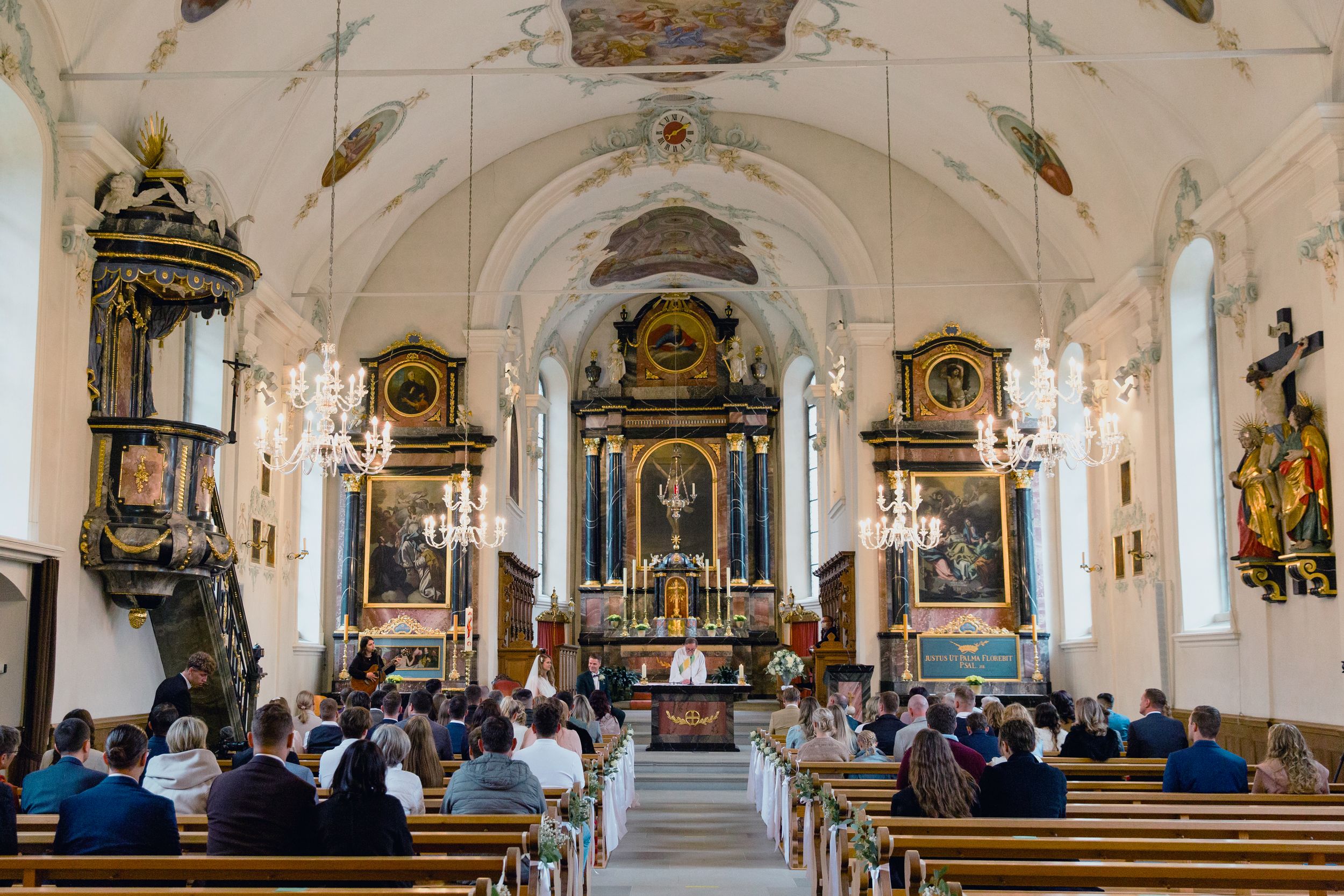 Wide interior view of an ornate Baroque church altar and nave during a wedding ceremony.