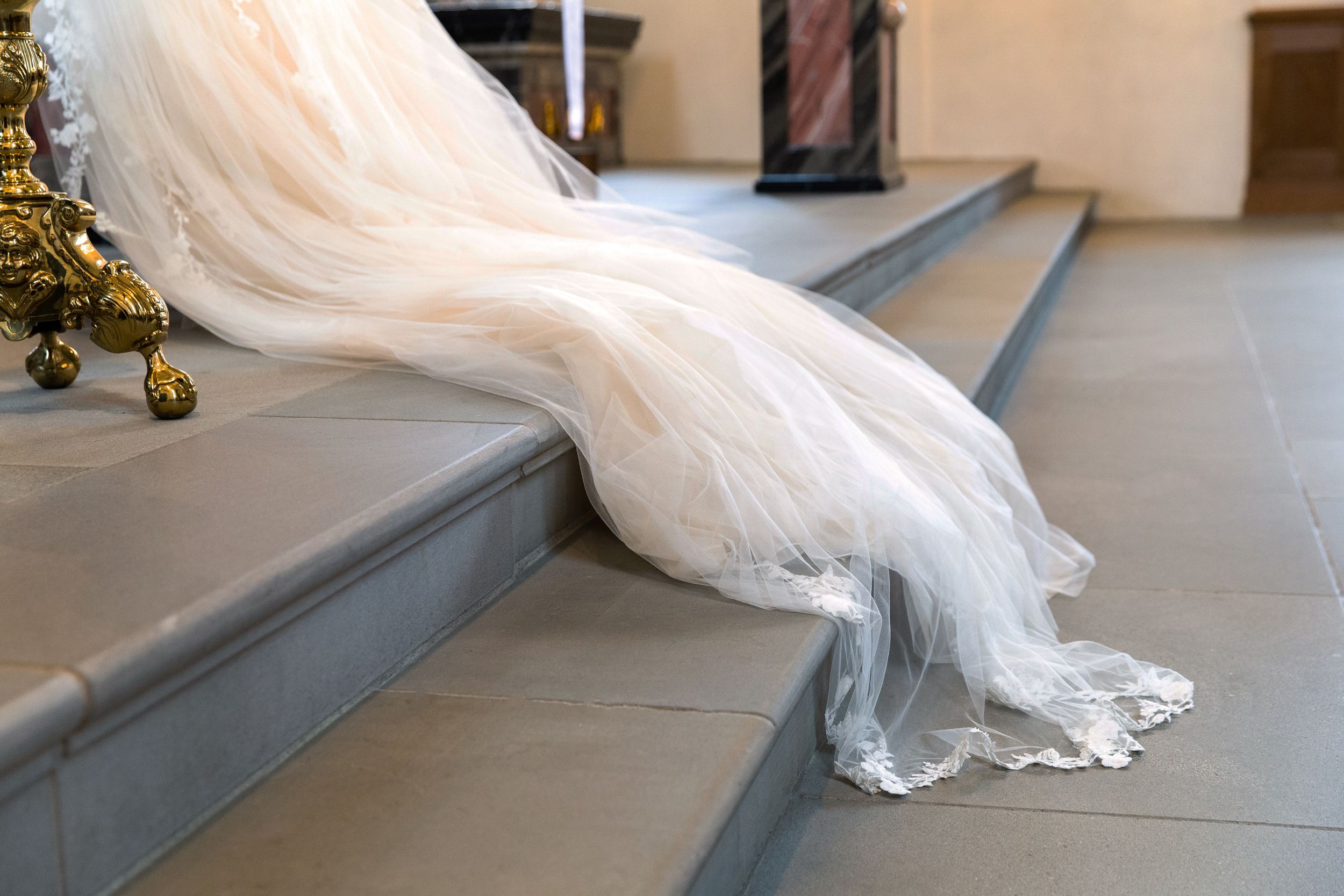 Close-up detail shot of a long, lace bridal veil trailing down the stone steps of a church altar.