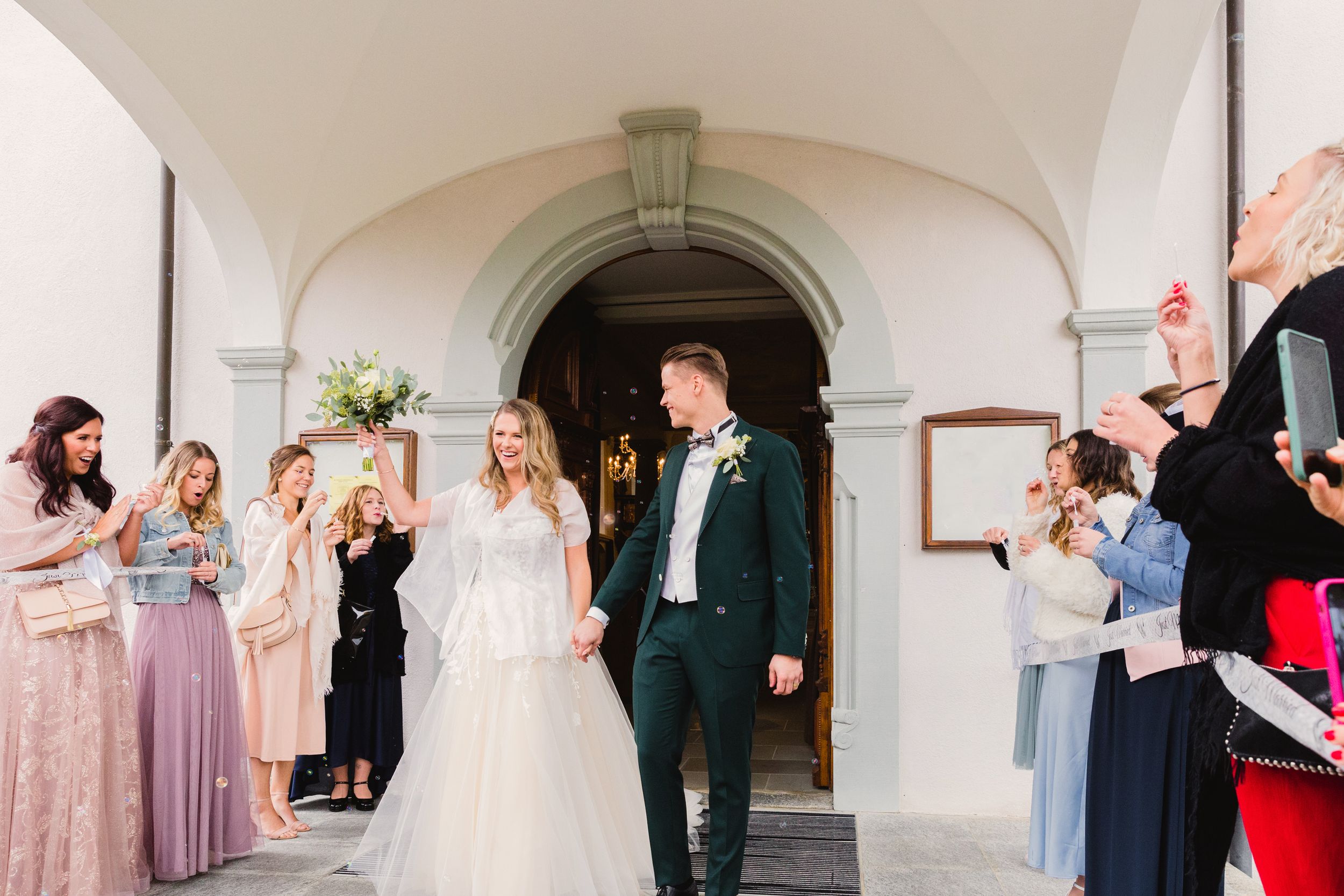 Bride and groom smiling and walking through a cheering crowd under a grand archway at their wedding.