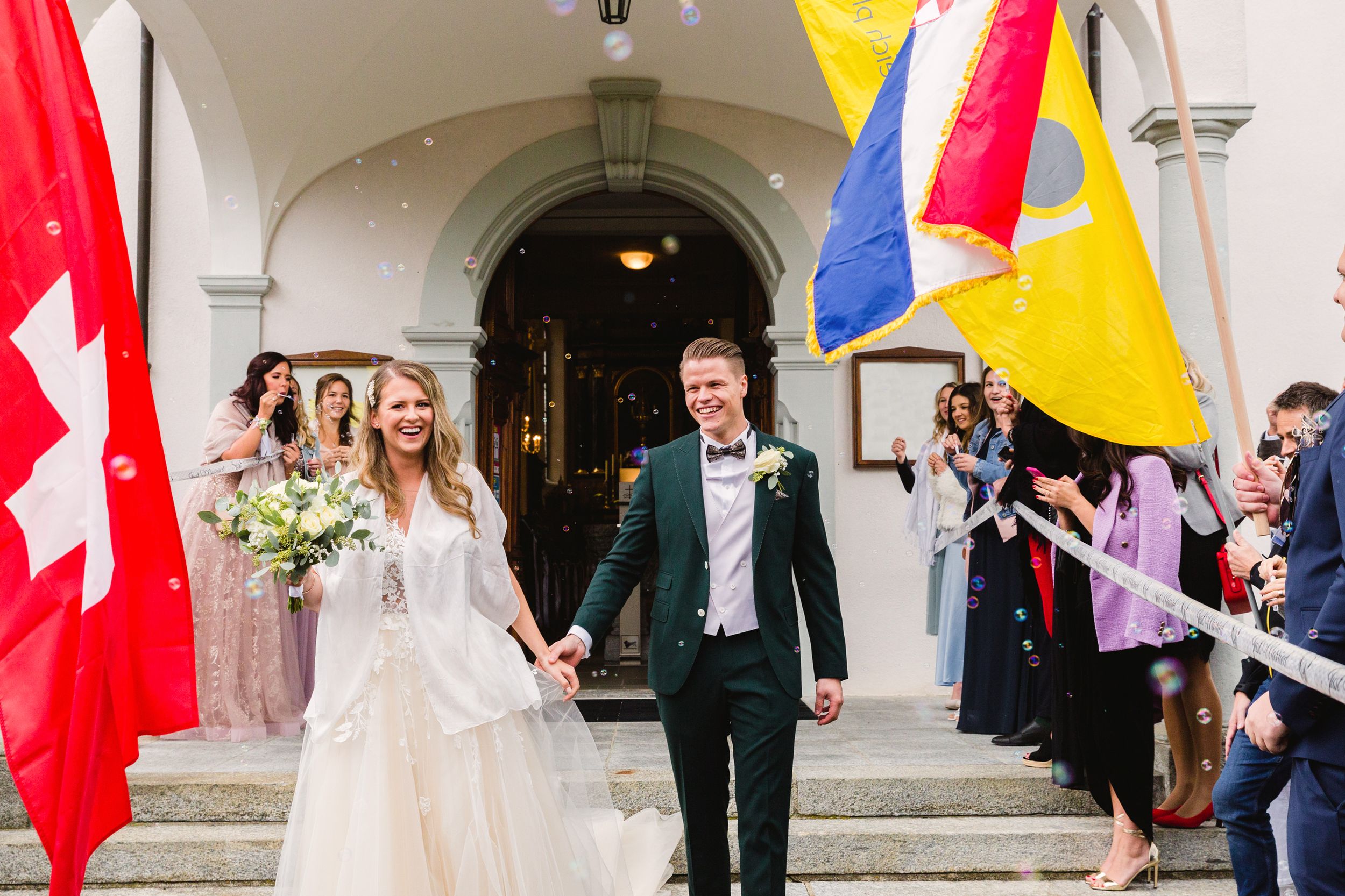 Newlywed couple walking out of a historic church entrance flanked by colorful Swiss flags.
