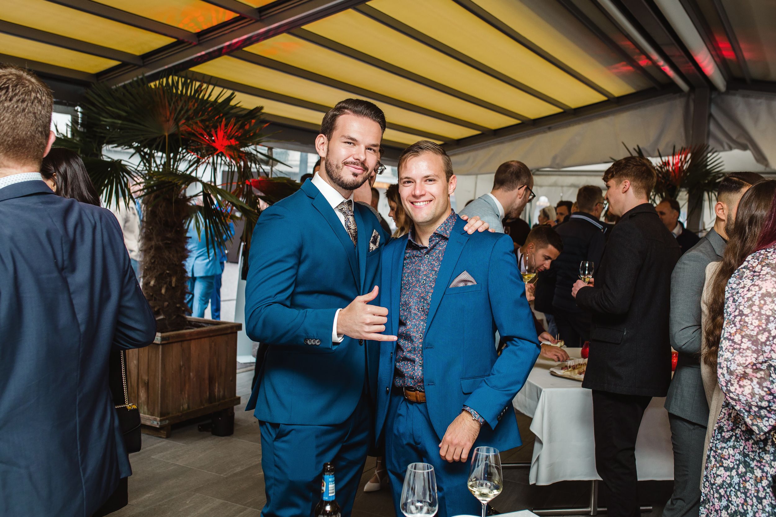 Two men in blue suits laughing and talking at an outdoor wedding cocktail hour.