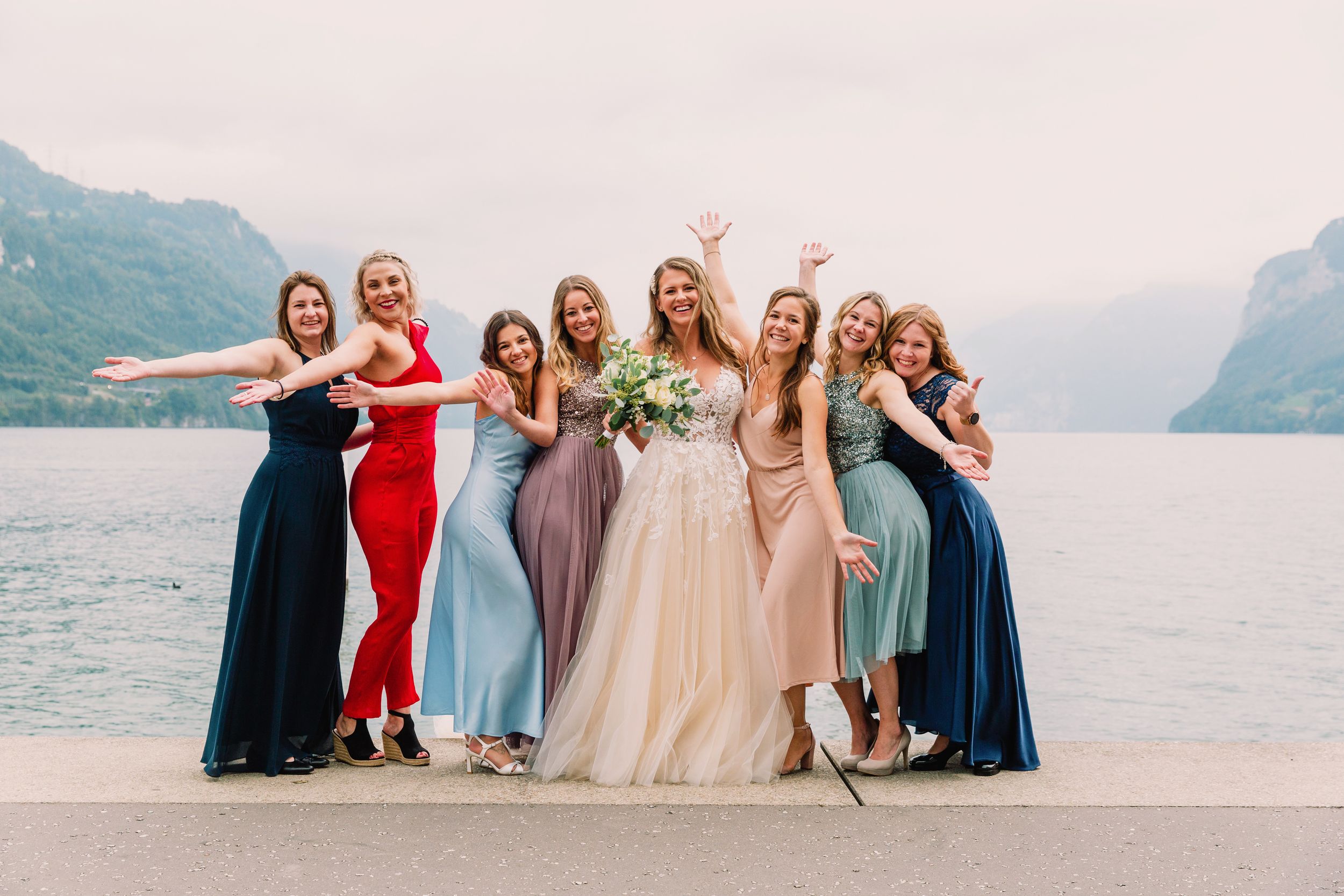 A group of bridesmaids in colorful dresses posing together on a lakeside terrace with a scenic mountain backdrop.