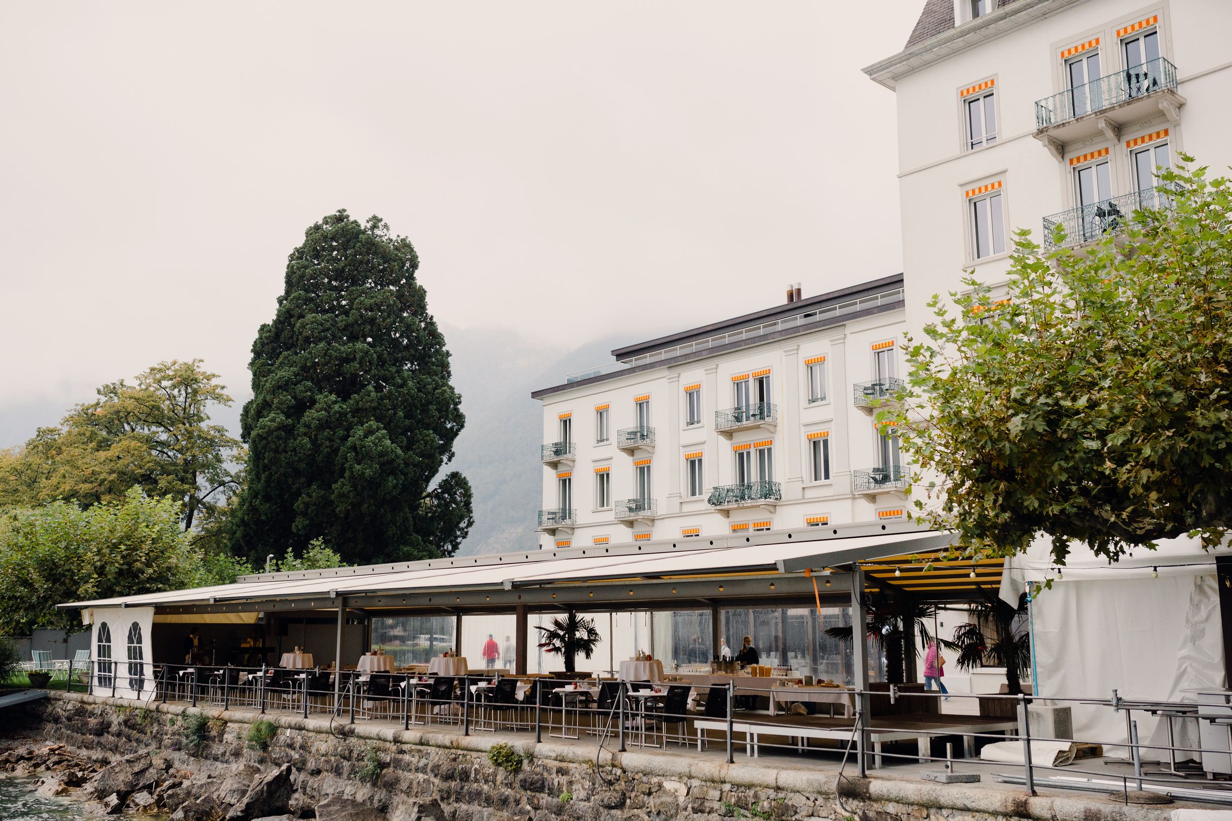 Wide shot of a historic white hotel and lakeside restaurant in Brunnen, Switzerland, surrounded by lush greenery.