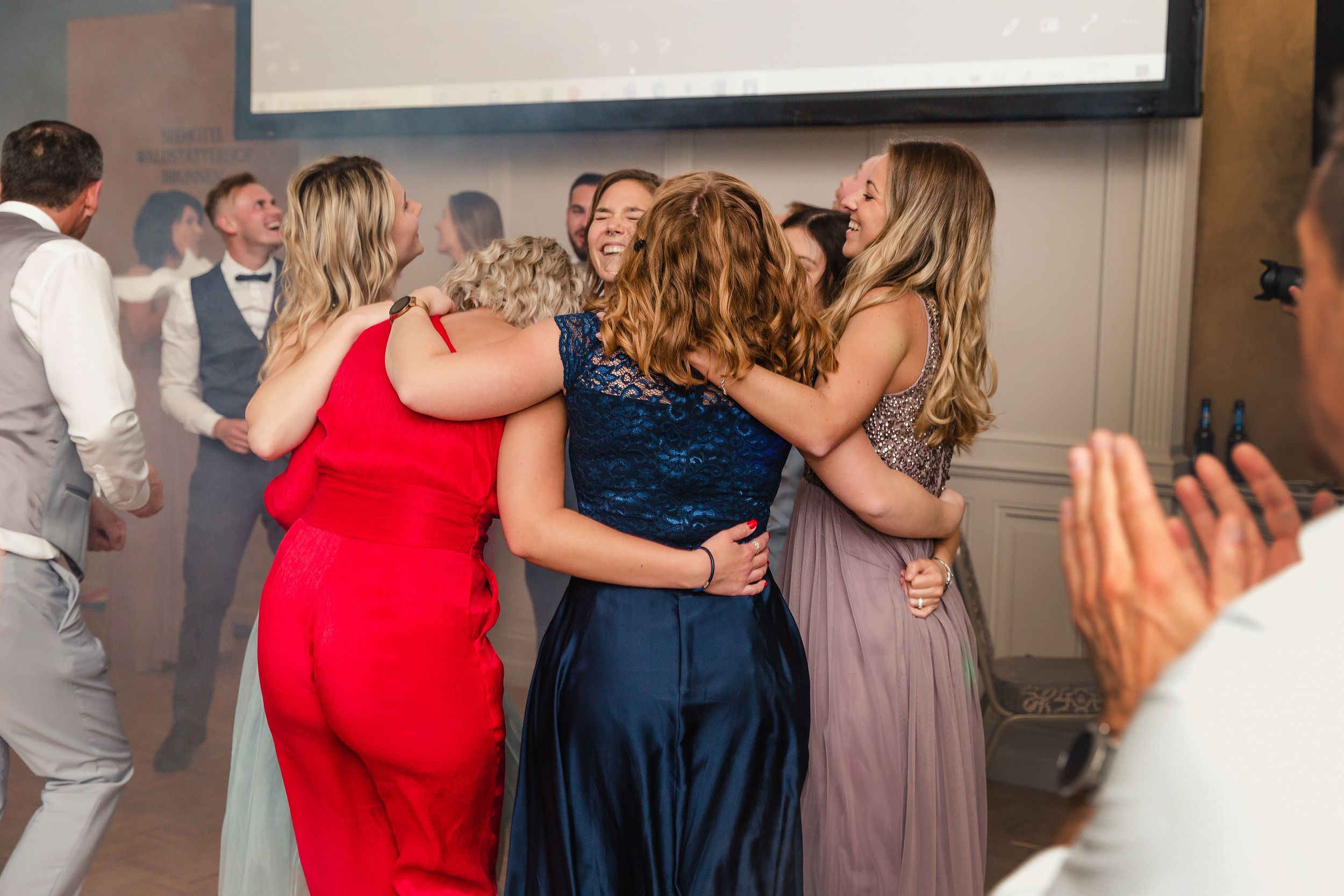 Candid photo of wedding guests in colorful dresses dancing and celebrating at an indoor reception.