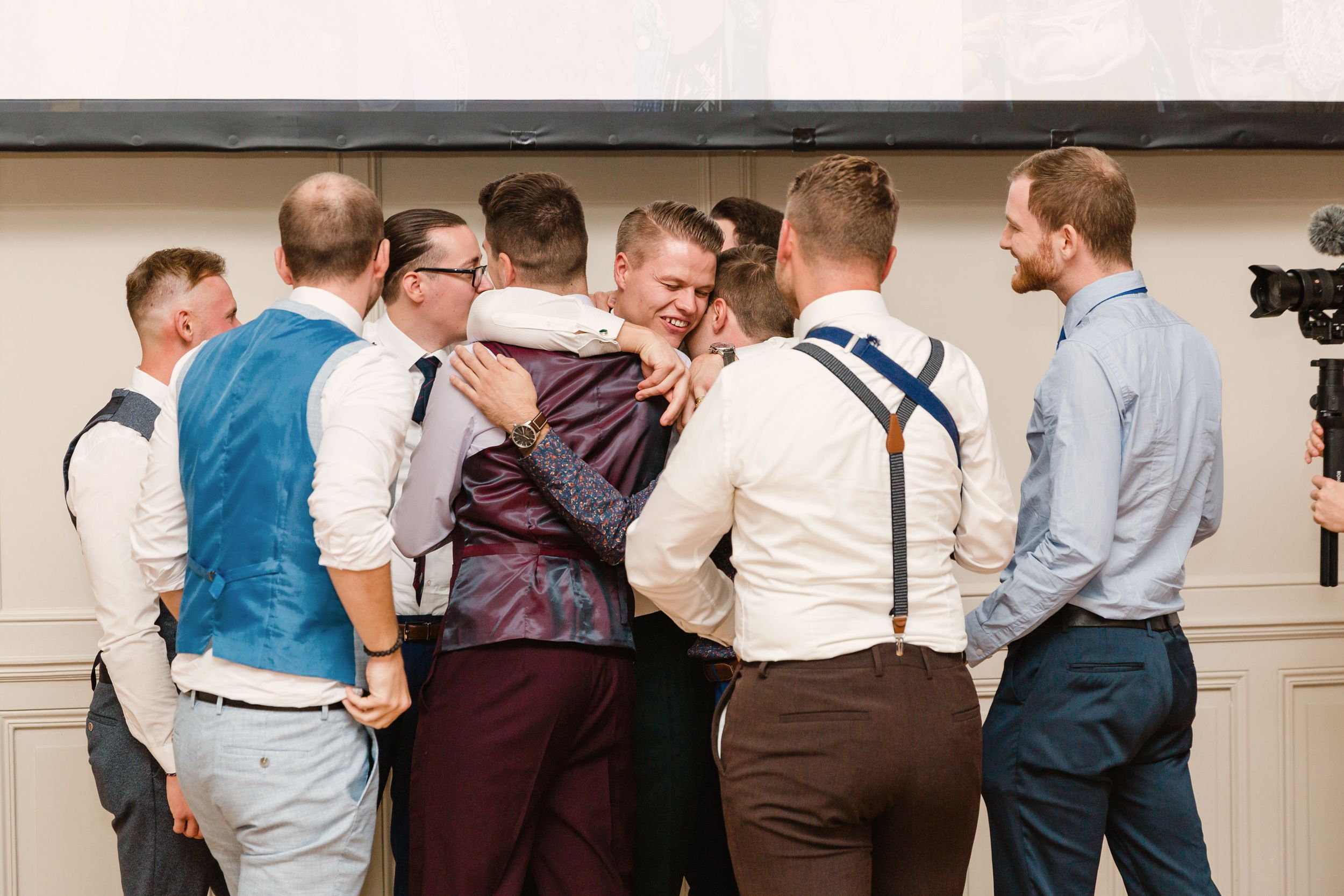 A group of groomsmen in vests and suspenders laughing and sharing a toast together.