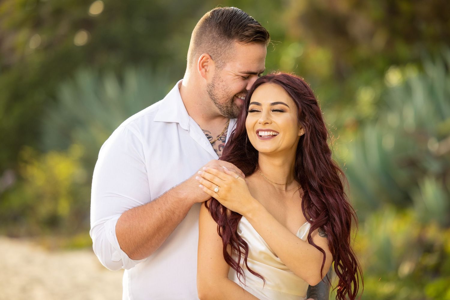 Romantic couple embracing outdoors with soft natural lighting and lush greenery in background.