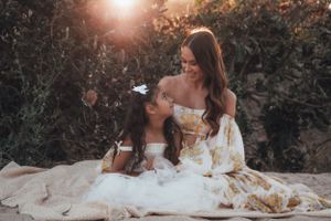Two figures in elegant dresses sit together in a dreamy sunlit garden setting with golden hour lighting.