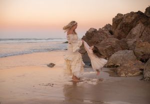 A person in a flowing cream dress twirls on a rocky beach at sunset.