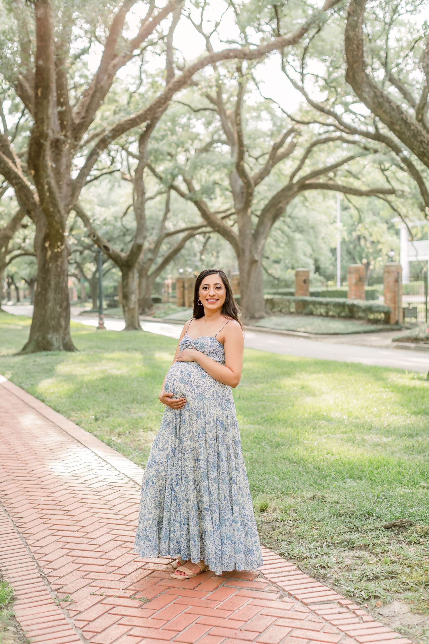 Expecting mother in flowing blue floral dress poses on brick pathway in serene park setting.