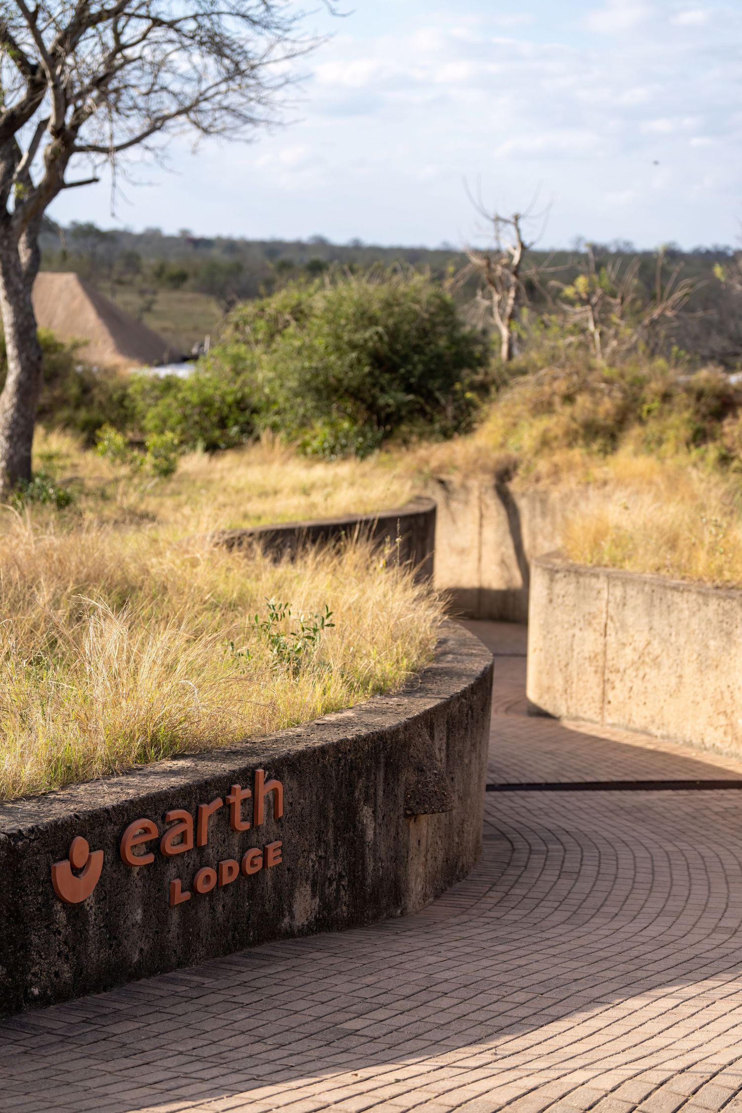 A curved brick pathway with 'earth lodge' text written on a retaining wall leads through an outdoor safari setting.