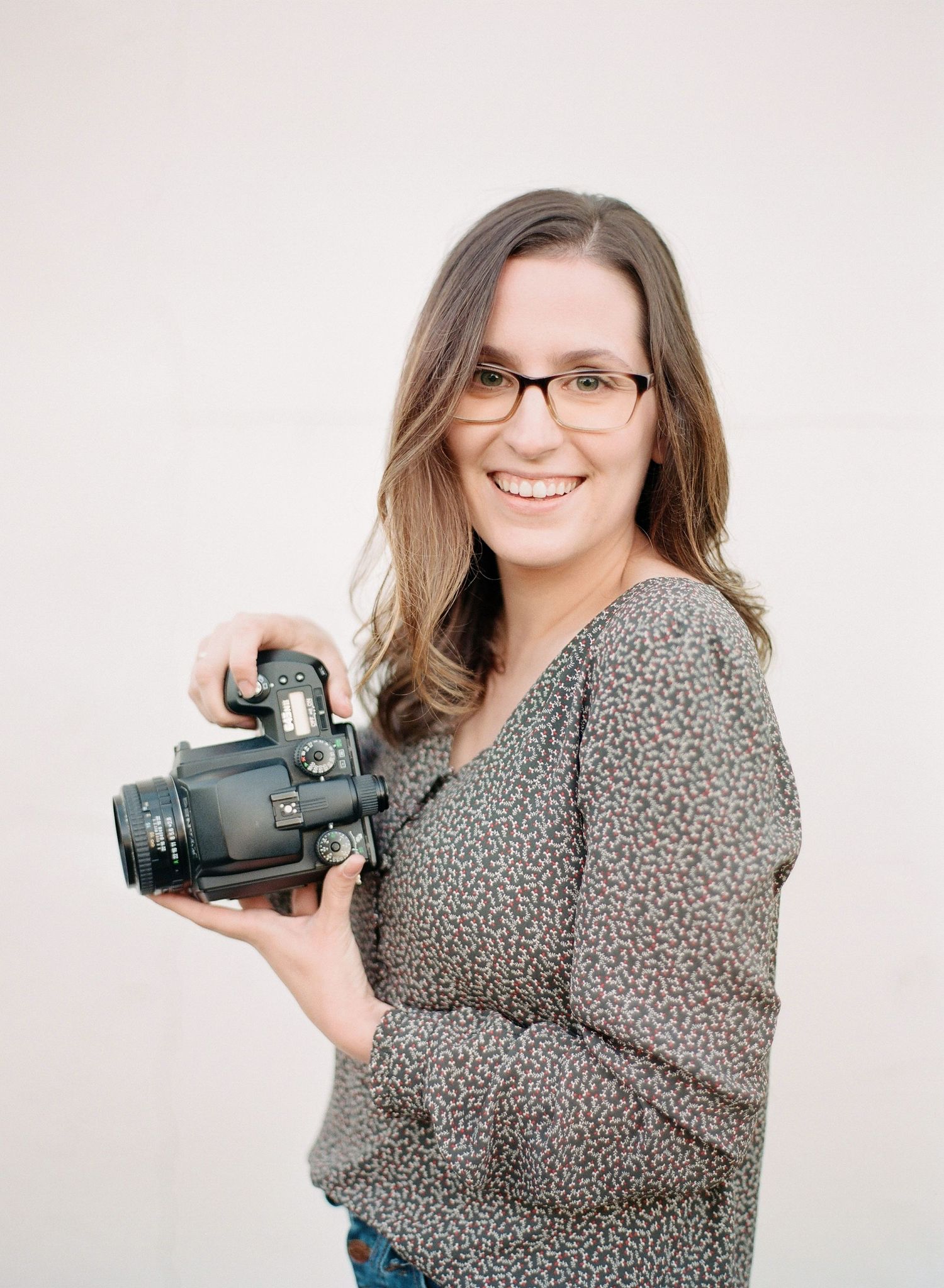 Professional photographer in a gray sweater holding a DSLR camera against a neutral background.