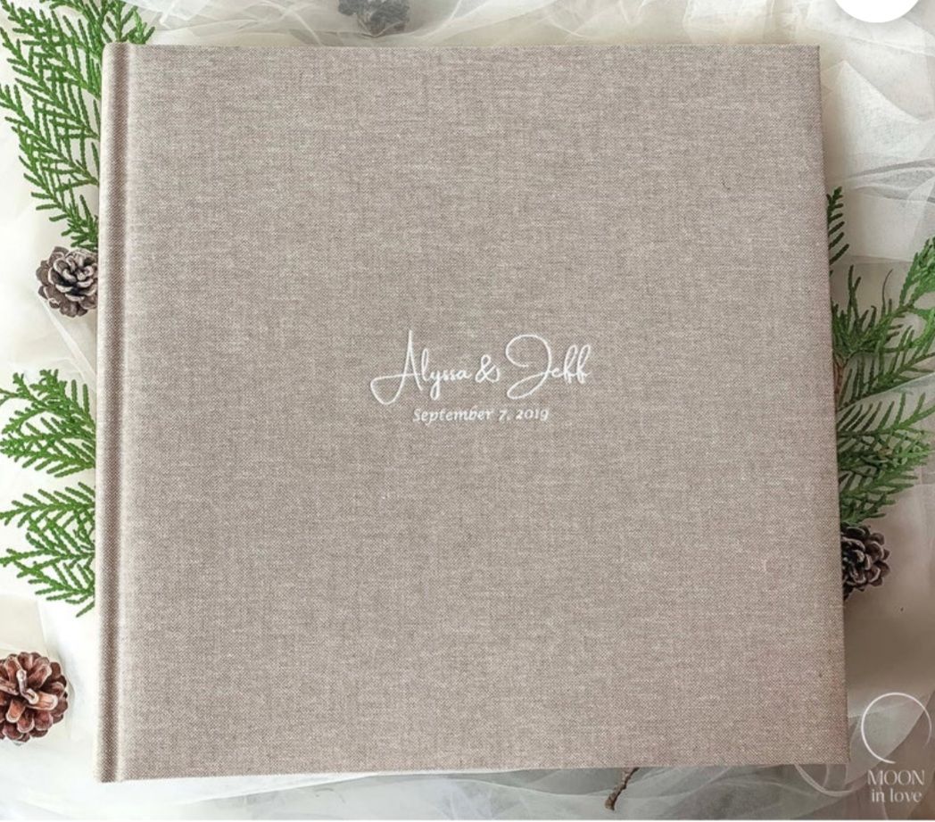 A minimalist linen wedding album with elegant script text lies among pine branches and pinecones.