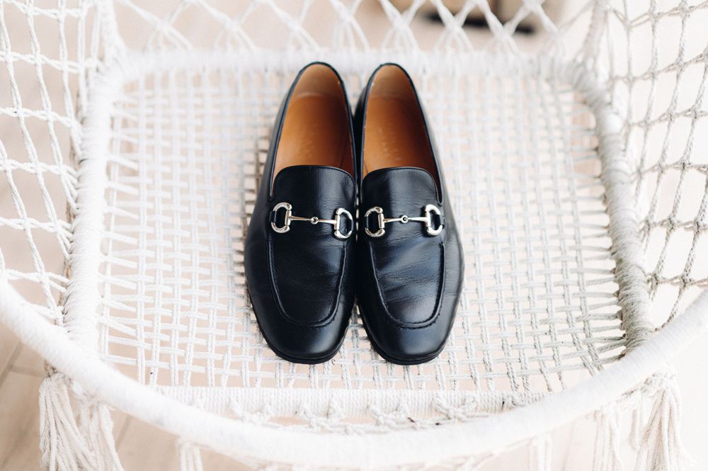 Black leather loafers with silver horsebit detail positioned on white wicker furniture.