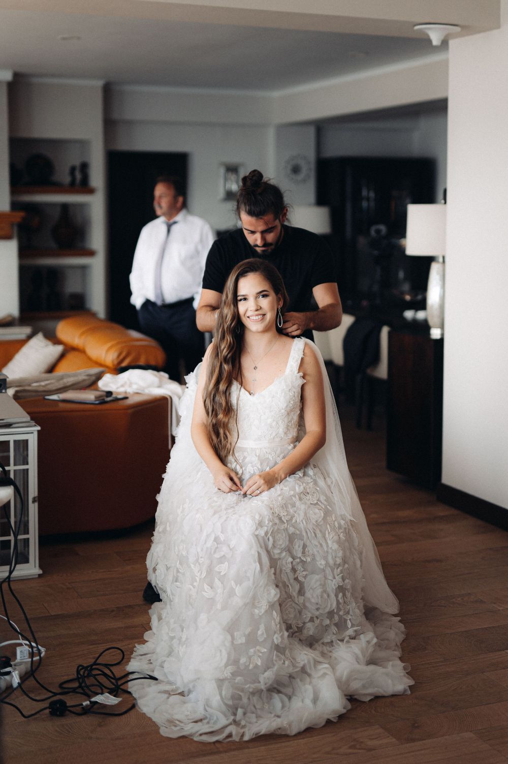 A bride sits in a lace wedding gown while getting her hair styled in preparation for the ceremony.