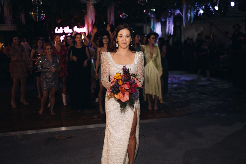 Elegant bride in sequined dress carrying vibrant orange and purple bouquet during evening reception.