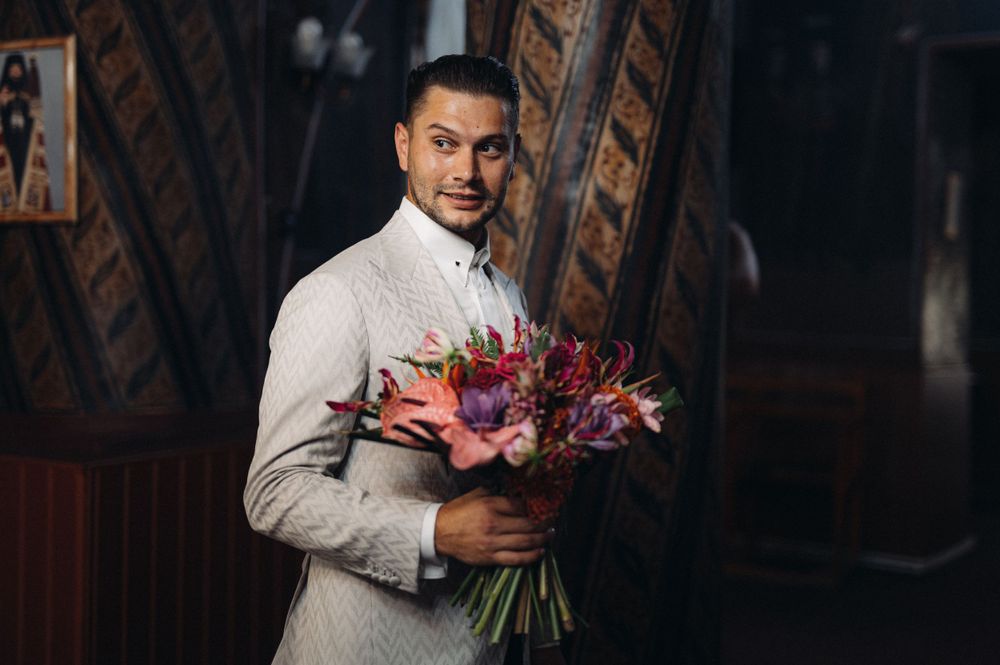 Groom holding colorful wedding bouquet in rustic wooden interior setting.