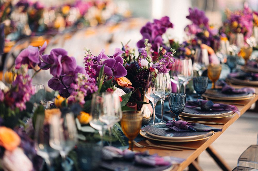 Elegant wedding table setting with purple floral centerpieces and dark plates lined up along a rustic wooden table.