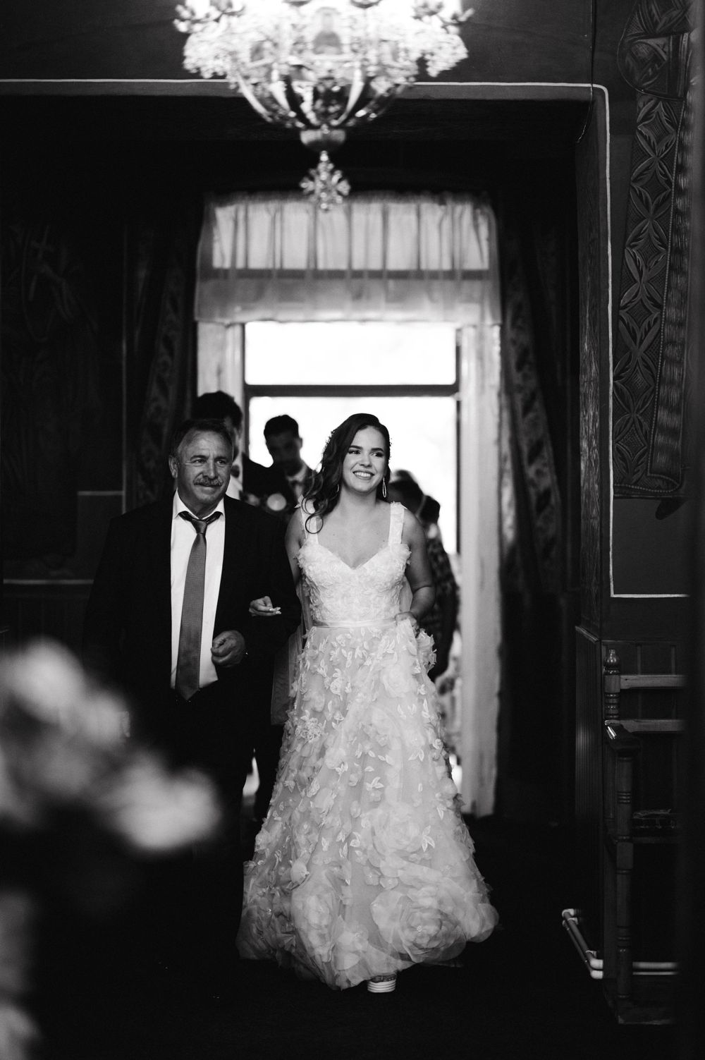 A black and white photo of a bride in a lace gown walking down a hallway with an escort under a crystal chandelier.