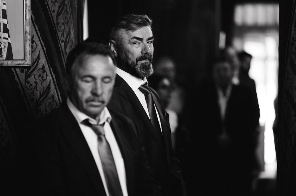 Two men in formal suits stand in a dimly lit hallway during a wedding ceremony.