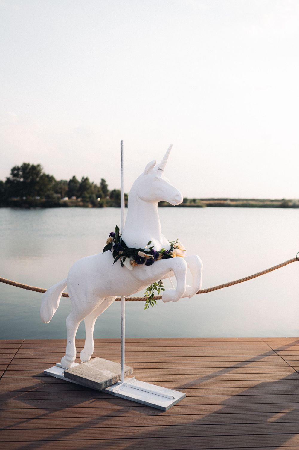 White decorative unicorn figure adorned with flowers displayed on wooden deck by waterfront.