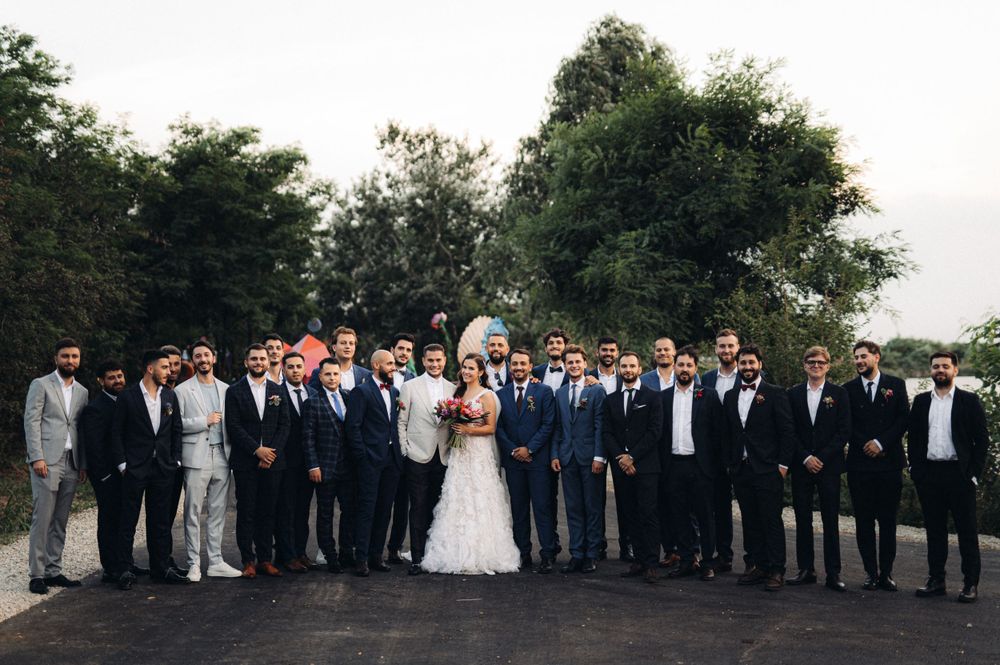 Wedding party and guests pose together outdoors in formal attire for a group photo.